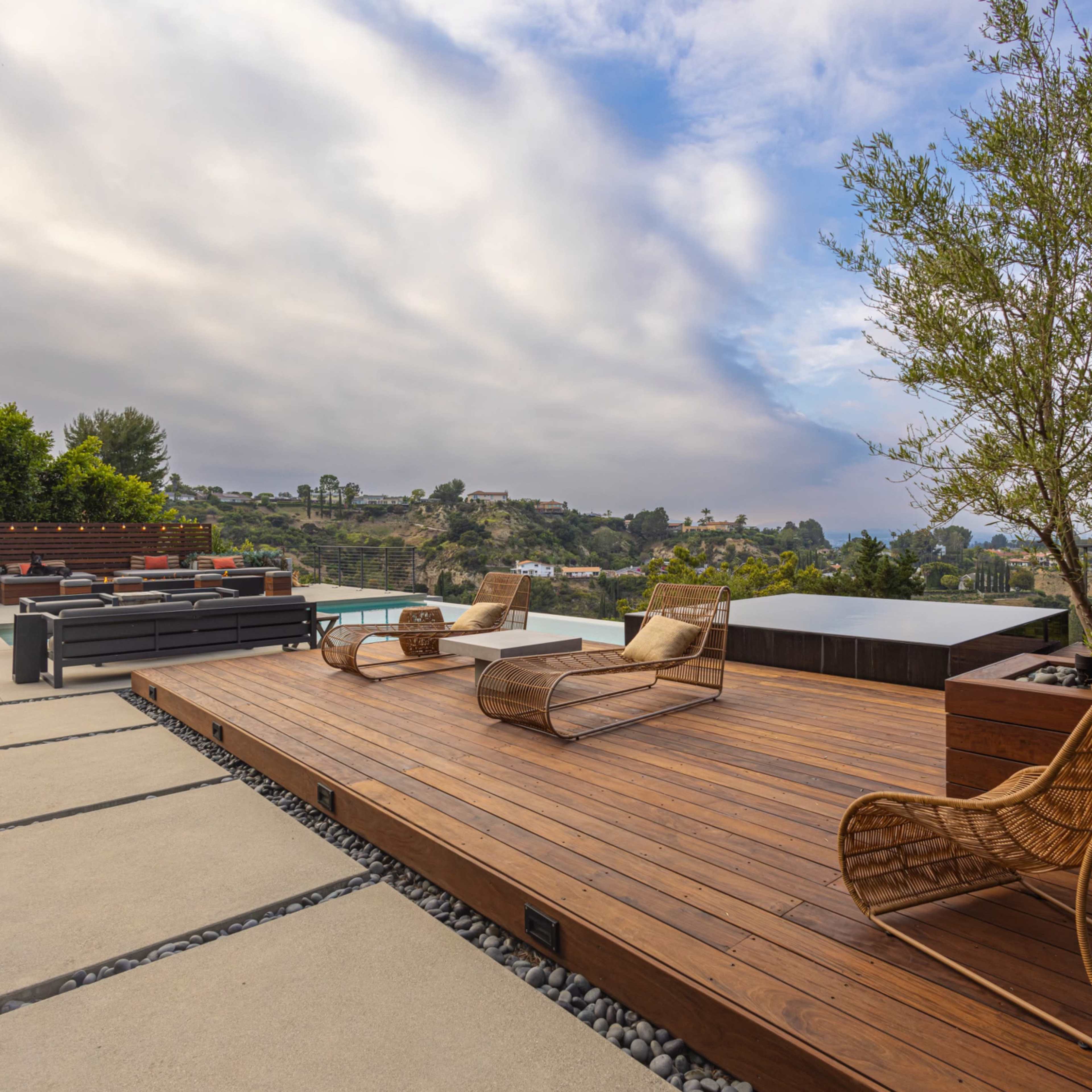 A wooden deck on a hillside with modern outdoor furniture and a view of a landscape under a cloudy sky.