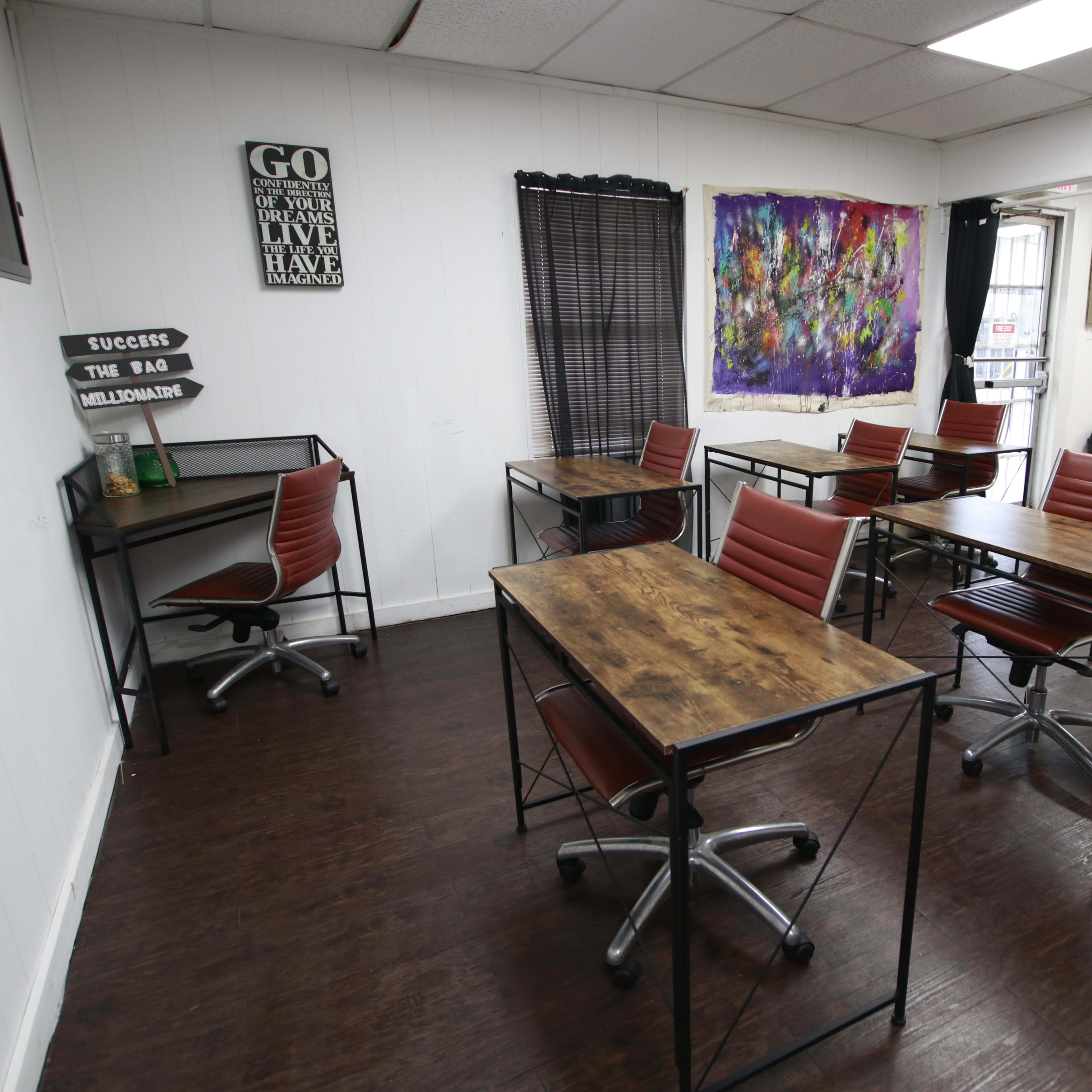 A study or workspace with multiple wooden desks and red chairs arranged in a bright, open room containing a kitchenette area and colorful wall art.