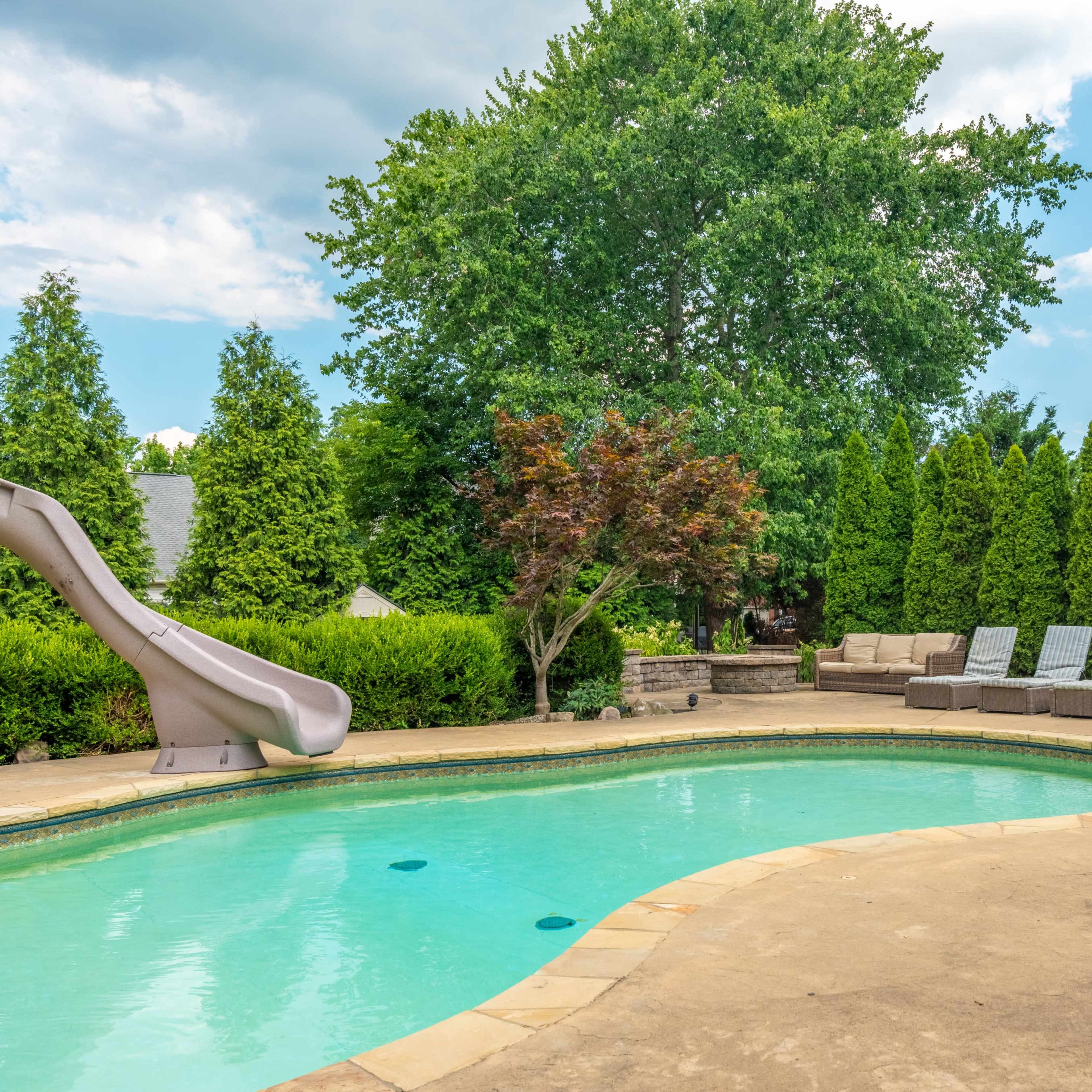 A backyard with a swimming pool, a water slide, lounge chairs, and a shaded seating area surrounded by tall green trees.