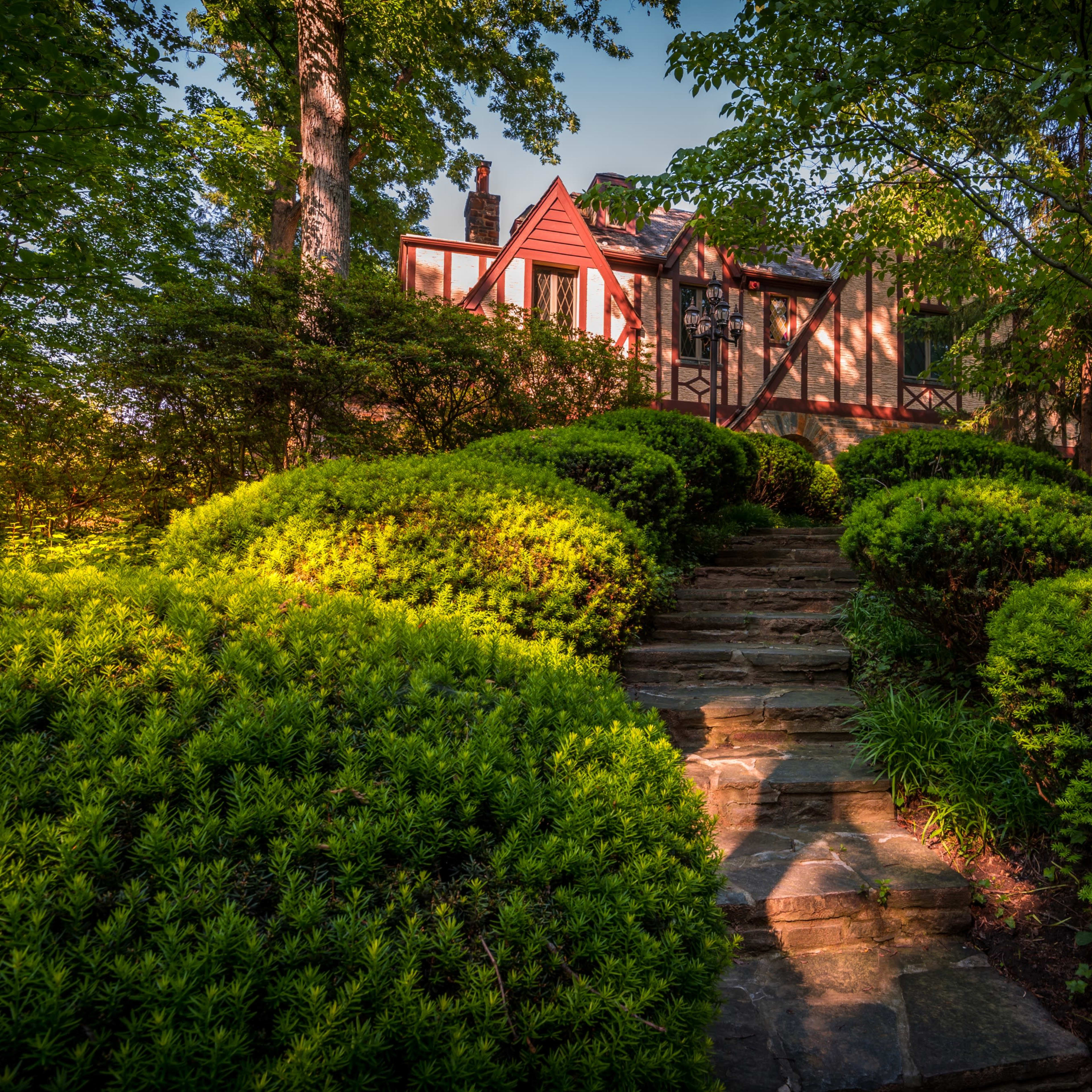 A stone pathway leads through lush green shrubs to a house partially obscured by trees.