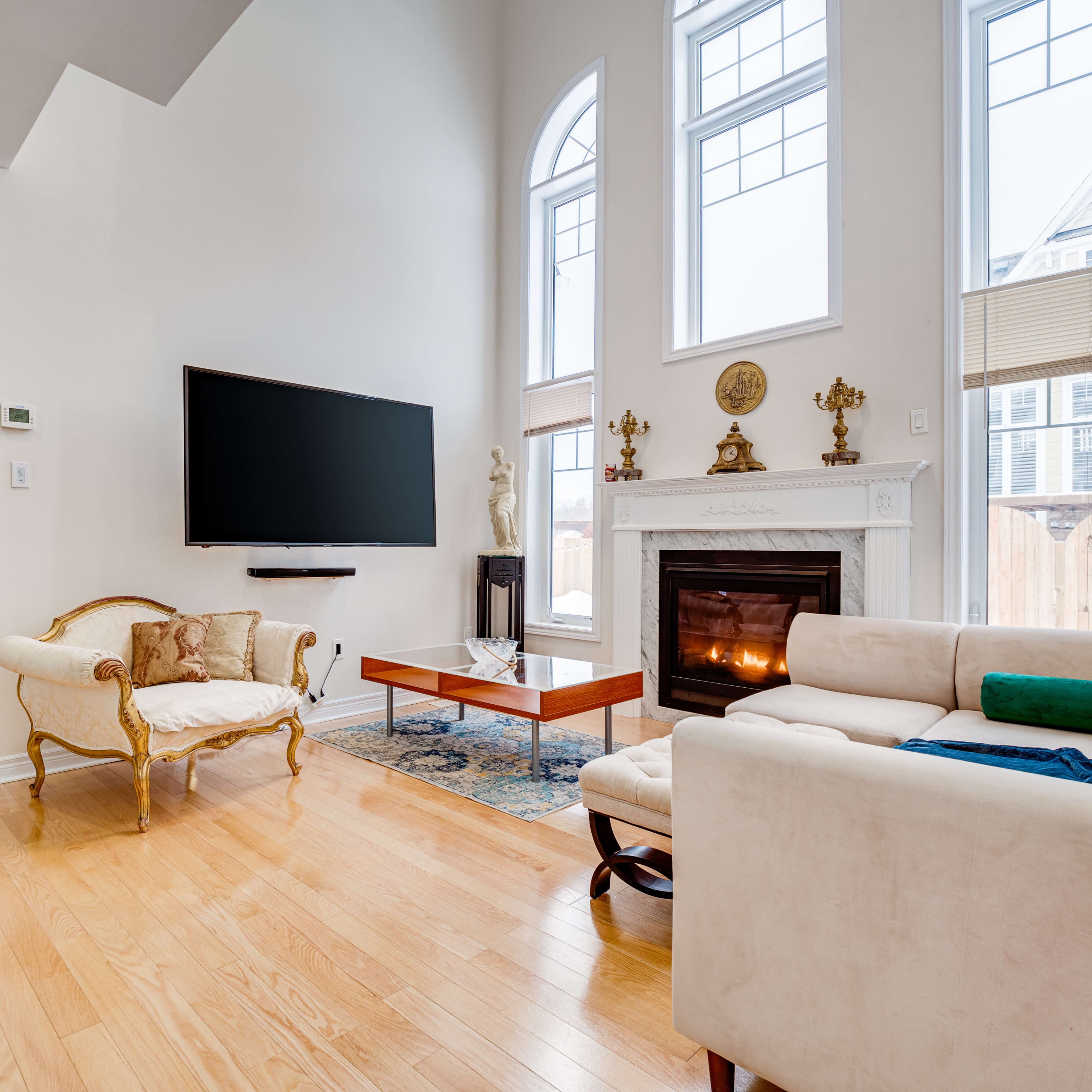 The scene features a living room with hardwood floors, a modern television mounted on the wall, a fireplace with a mantel, and a mix of contemporary and traditional furniture.