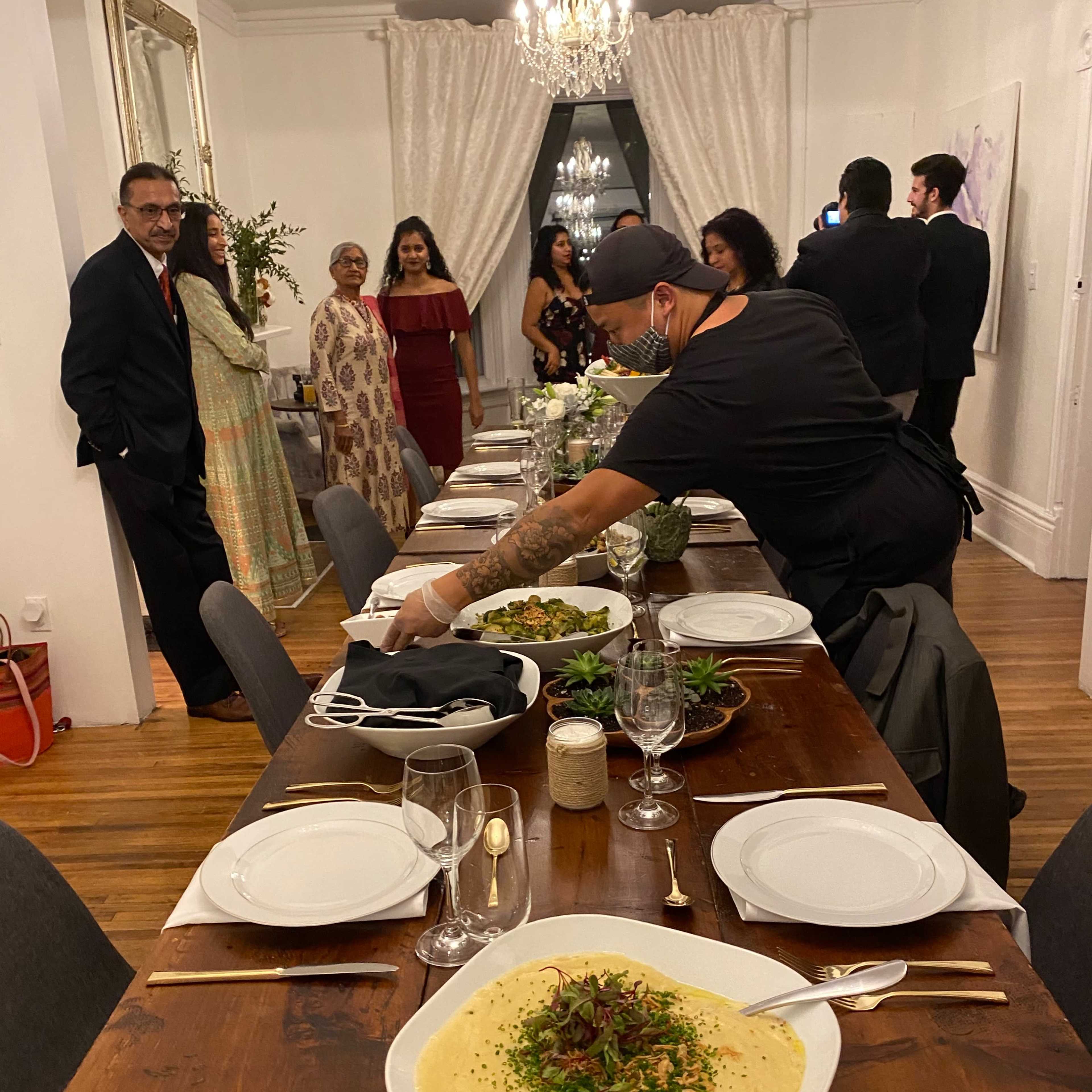 A long dining table is set for a gathering, adorned with dishes and surrounded by guests preparing for the meal.