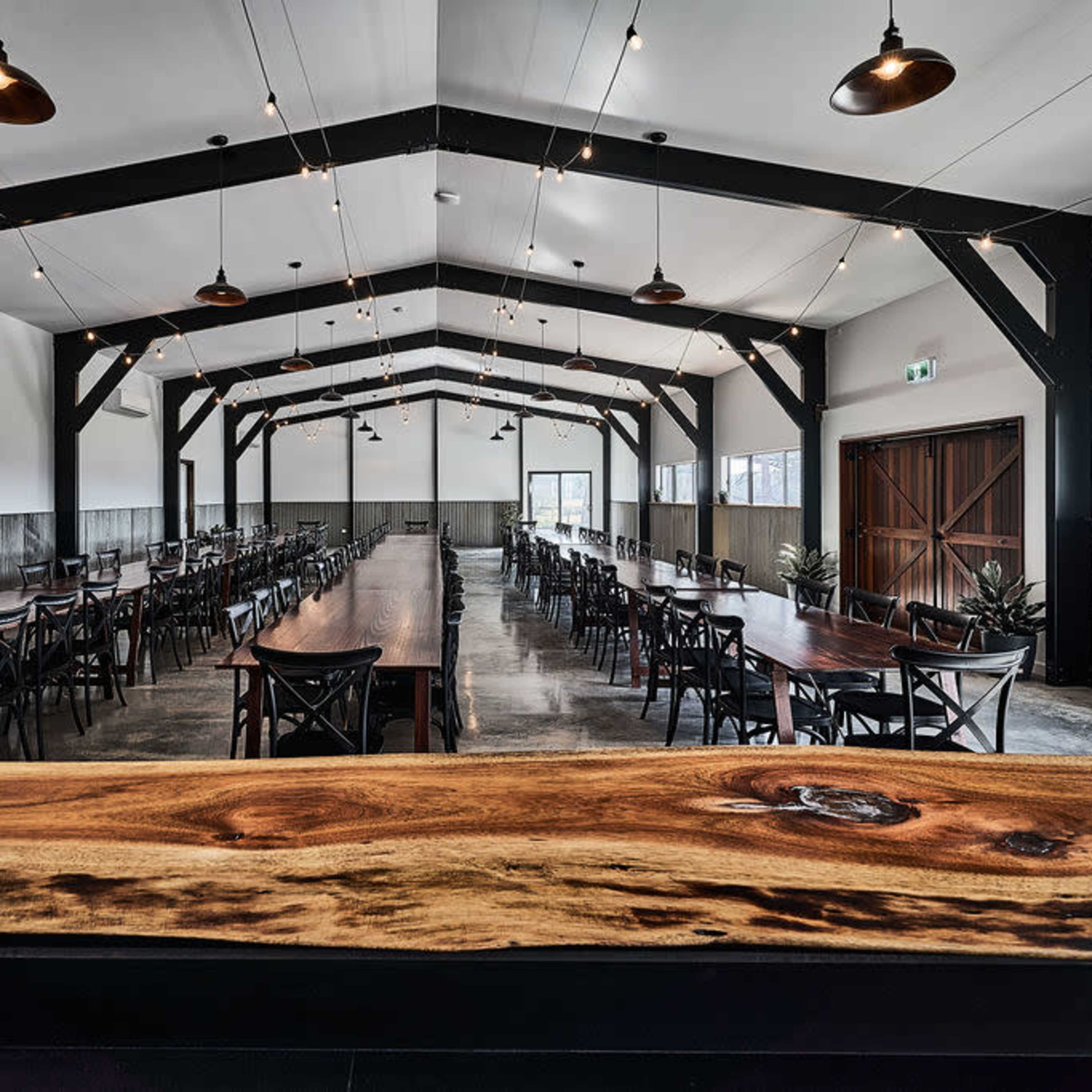 A large dining hall with long tables arranged in rows under a high ceiling supported by wooden beams, illuminated by overhead lights.