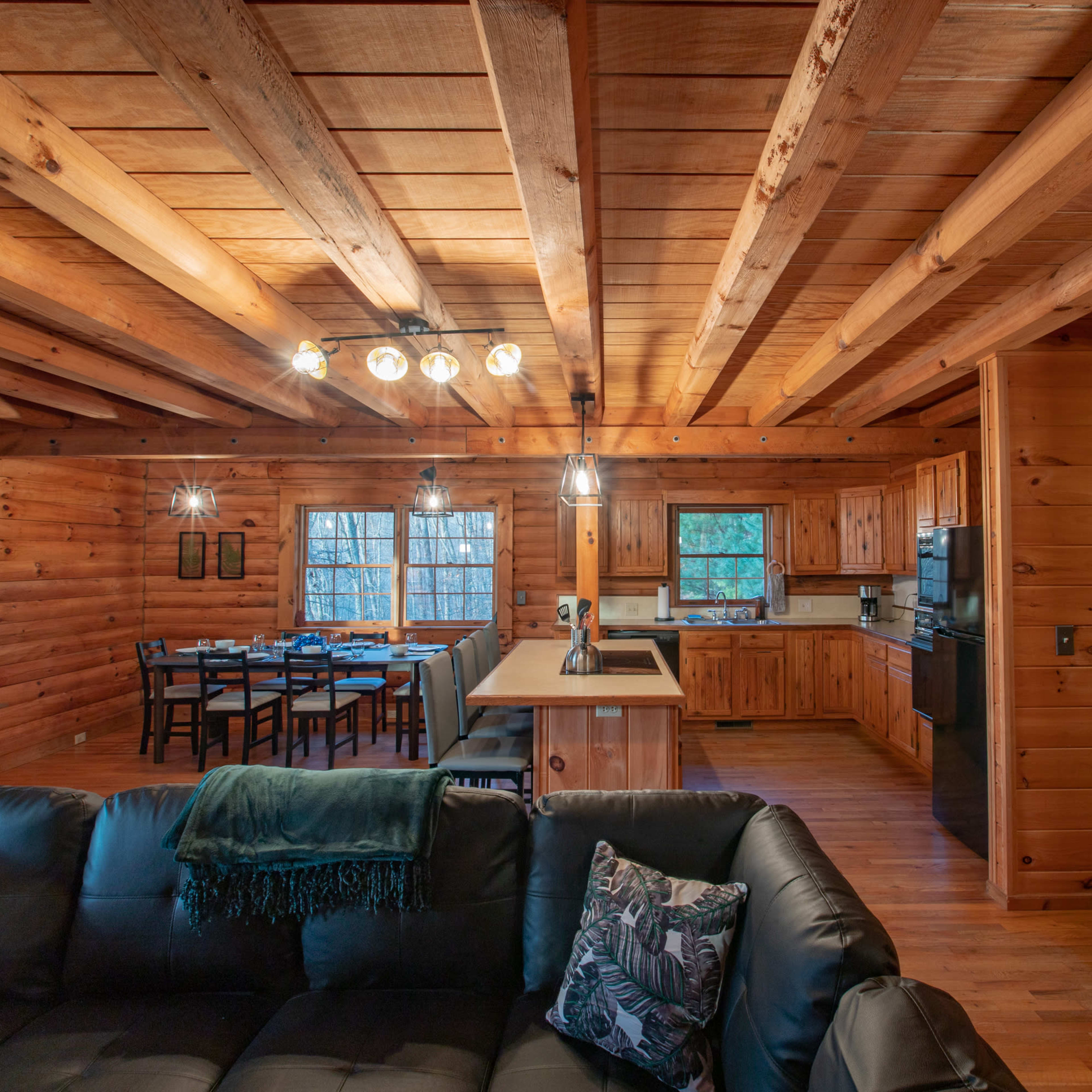 The interior of a wooden cabin features a cozy living area with a black sofa, an adjoining kitchen, and a dining area with a table set for six.