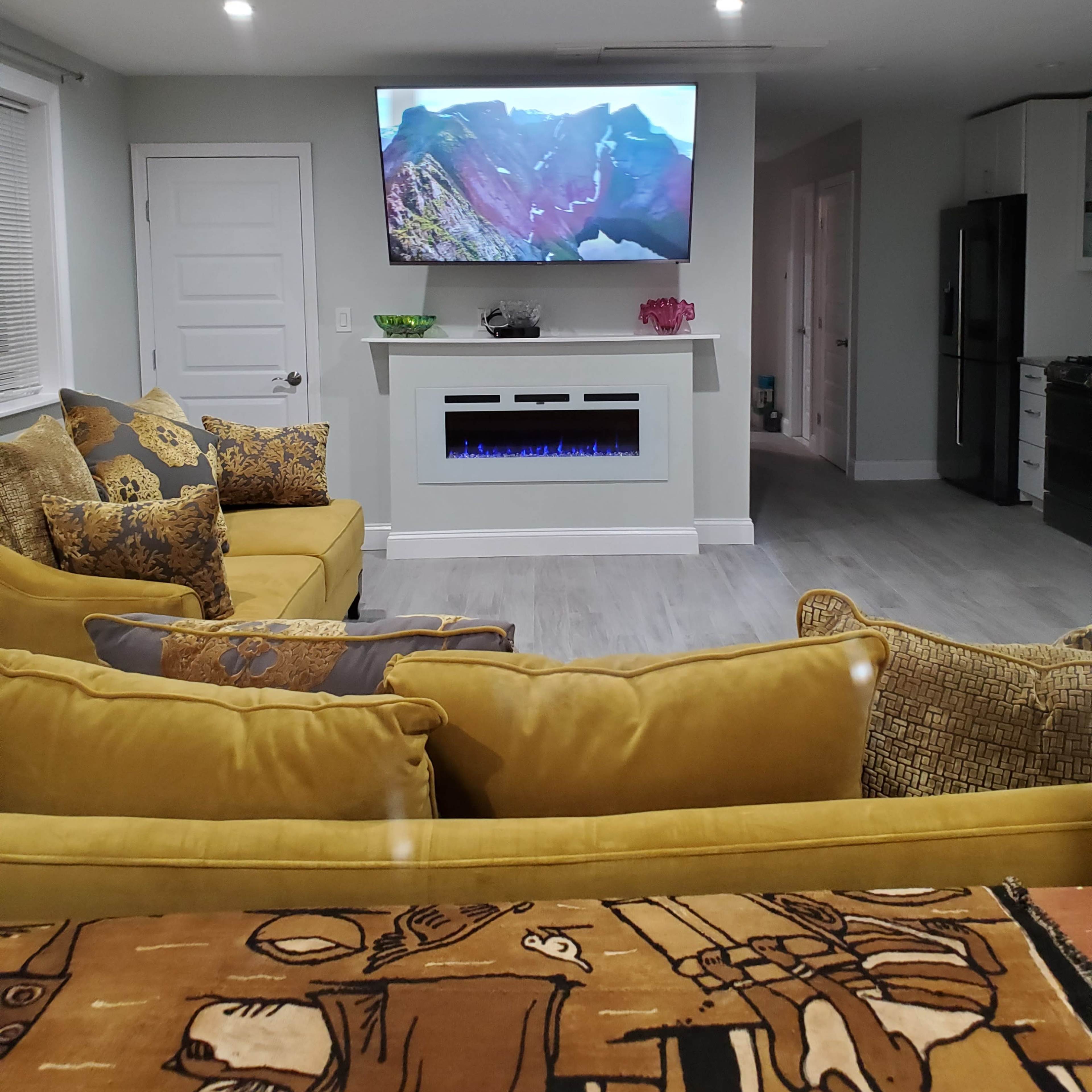 A modern living room features a yellow sectional sofa, a wall-mounted television displaying a mountain landscape, and an electric fireplace beneath the TV.