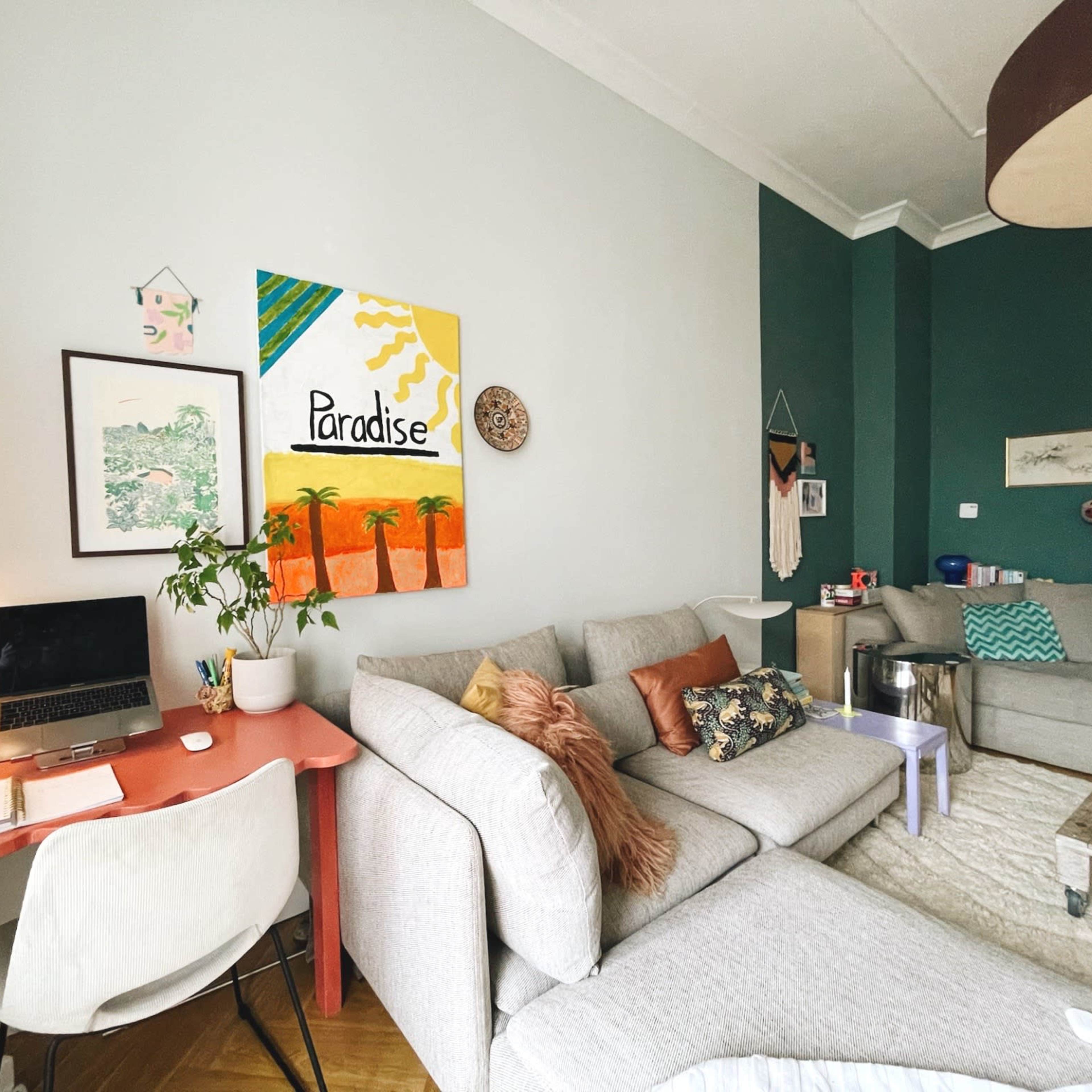 The living room features a light gray sectional sofa, a coral desk with a laptop, and vibrant artwork, including a painting titled "Paradise" on the wall.