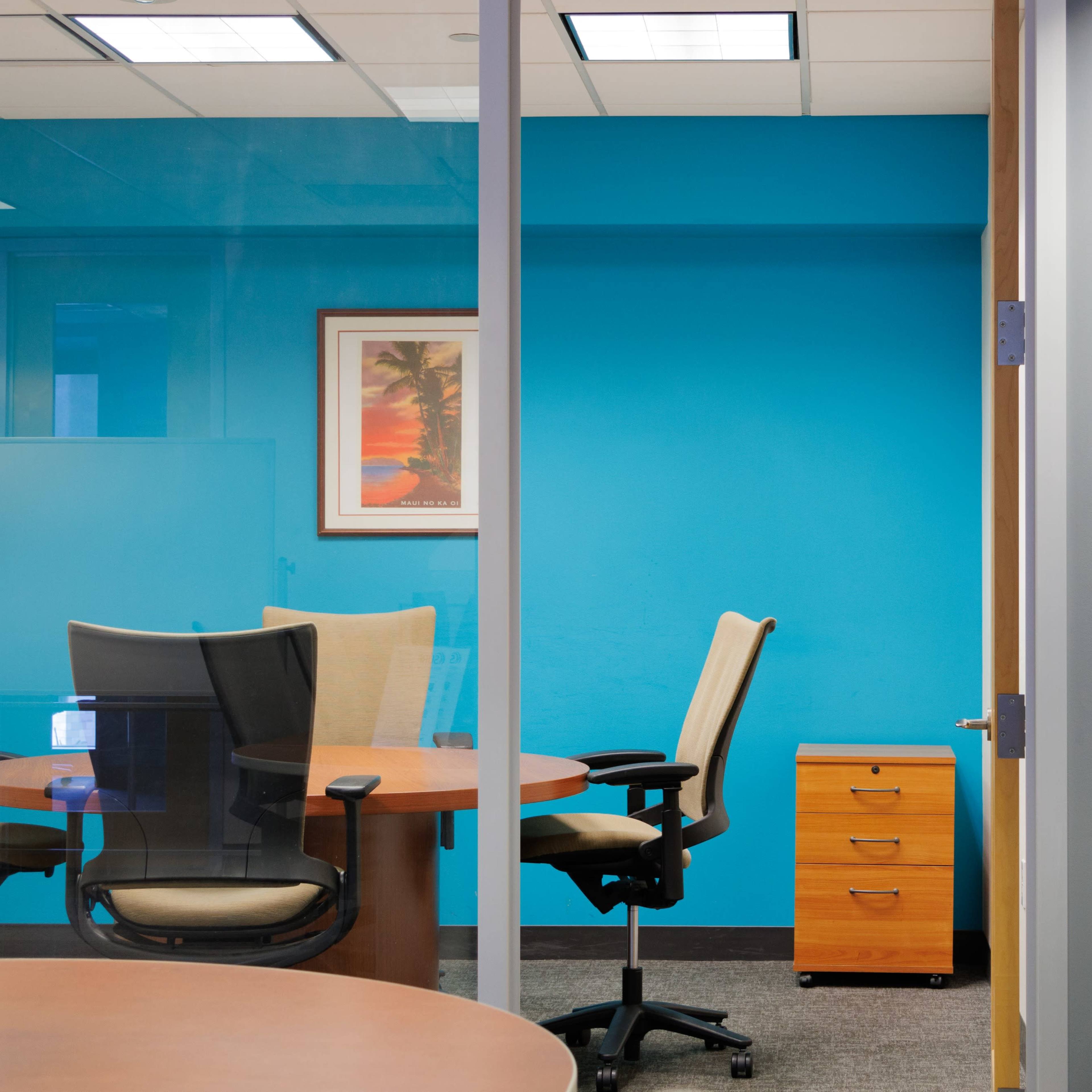 A modern office space features a glass partition, a blue accent wall, round tables with chairs, a wooden cabinet, and a potted plant.