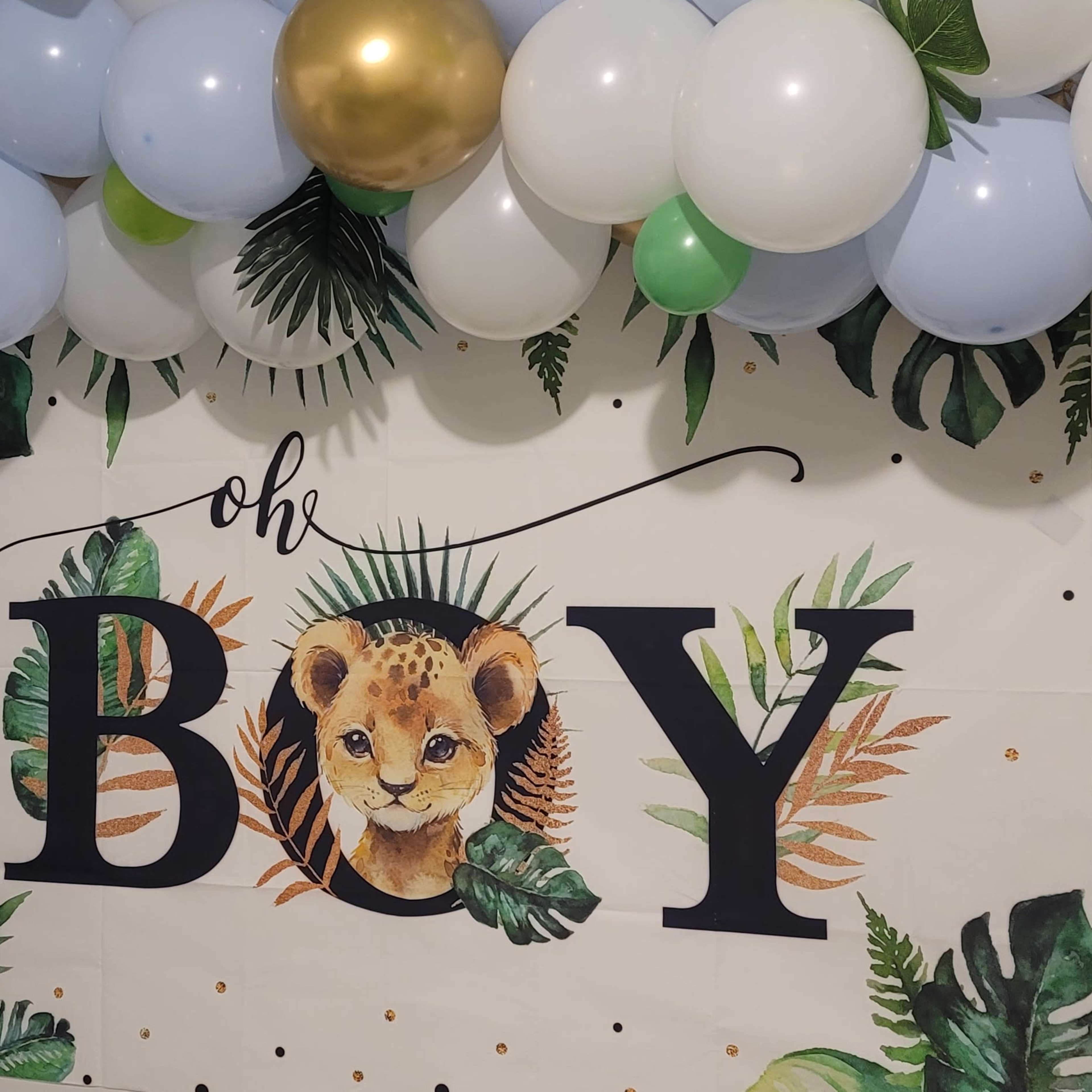 A decorated wall with a banner reading "oh BOY" featuring a lion cub and surrounded by a variety of balloons in green, blue, white, and gold.