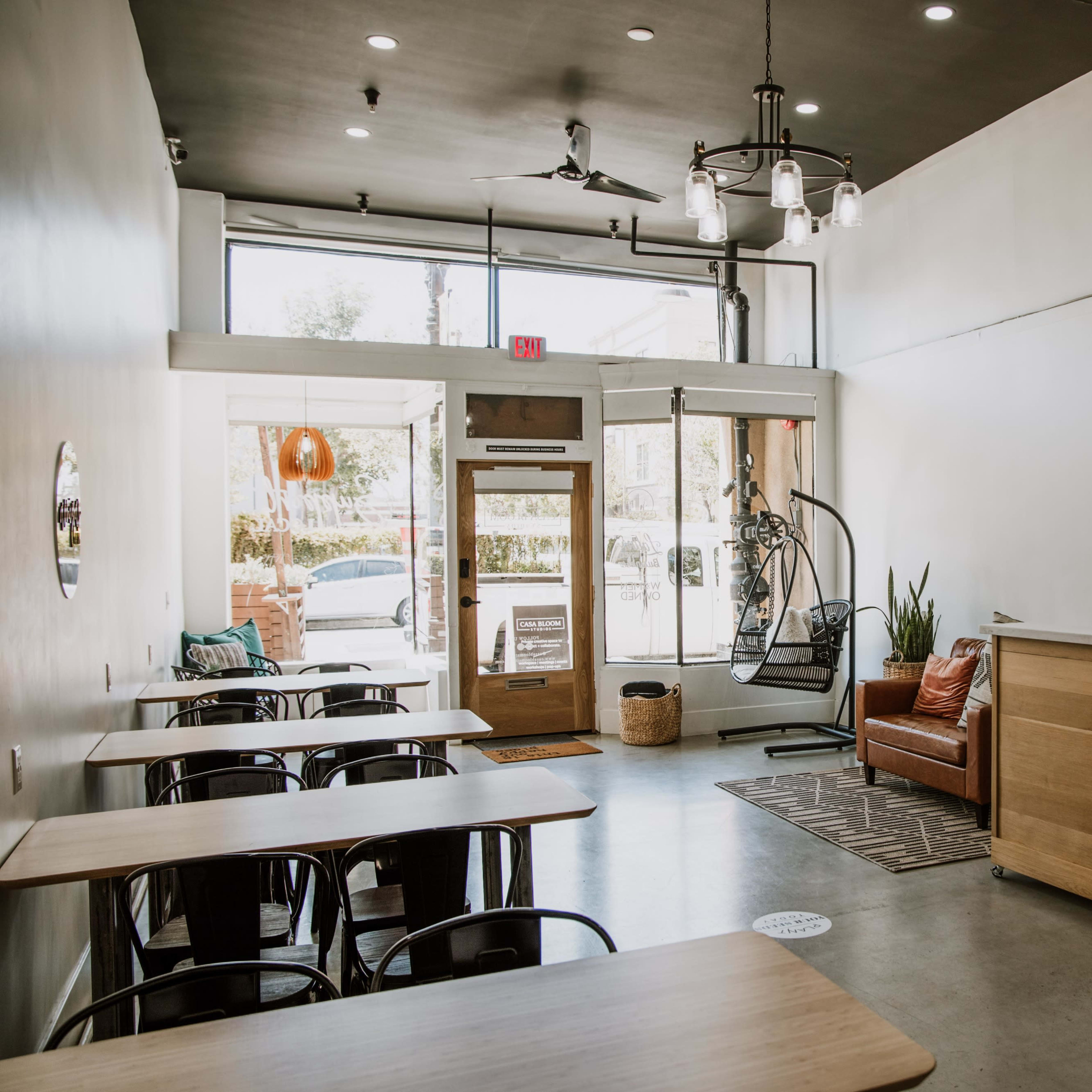 A modern cafe interior with a series of wooden tables, a seating area featuring a leather couch, and large windows letting in natural light.