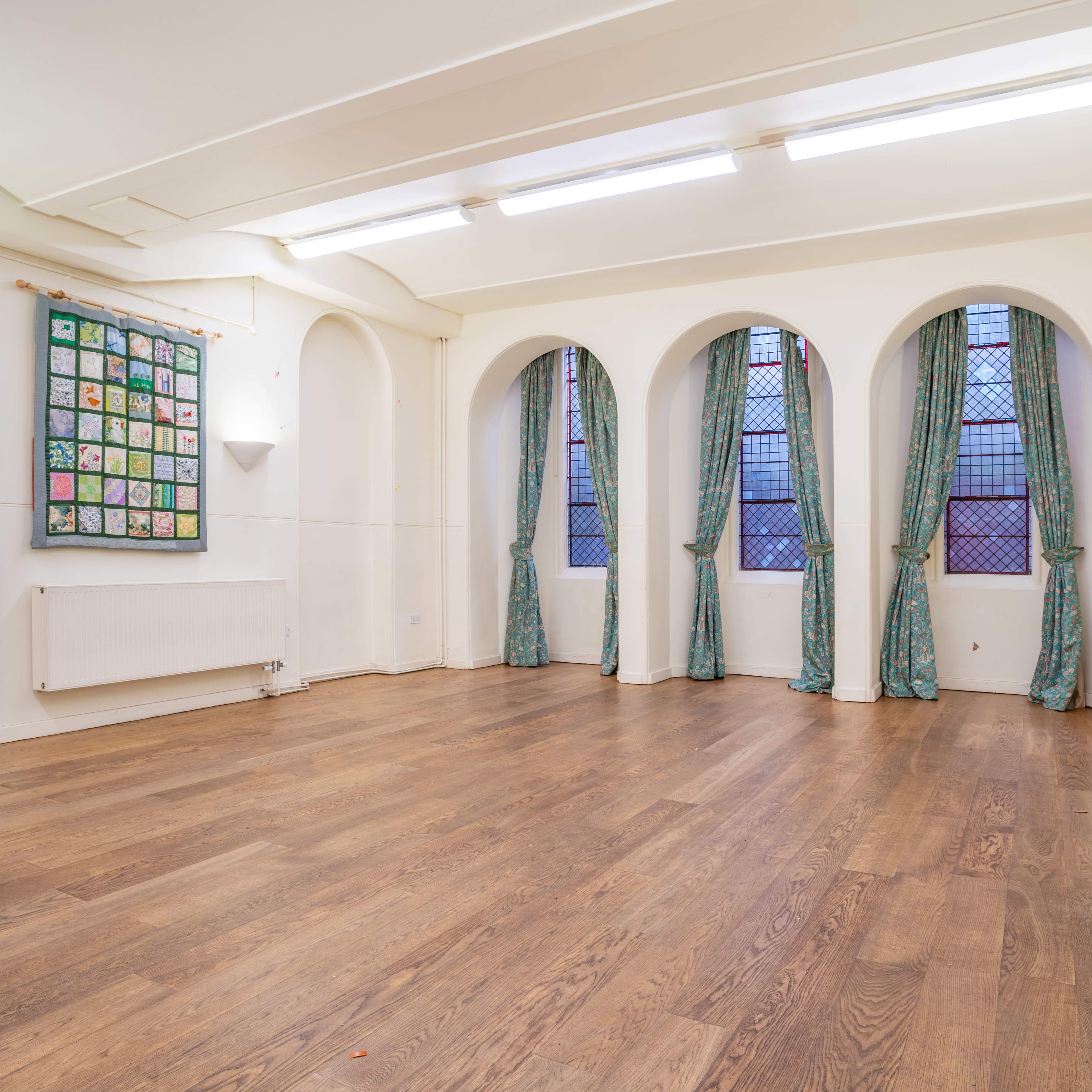A spacious, empty room with wooden flooring, white walls, and large arched windows draped with green curtains, alongside a wall adorned with a colorful display of framed artwork.