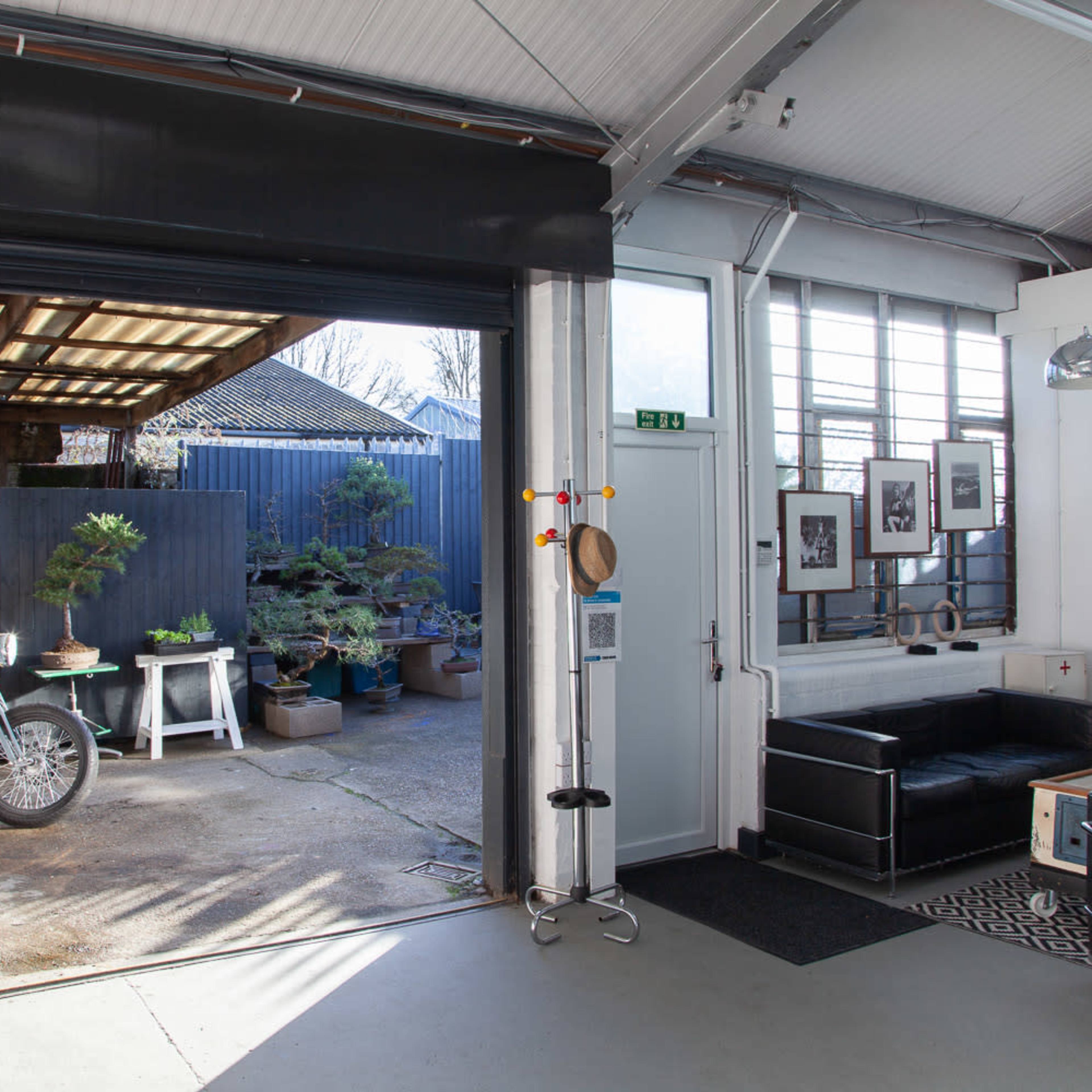 A spacious garage with an open door revealing a motorcycle and potted plants in the driveway, alongside a modern interior featuring a black sofa and decor.