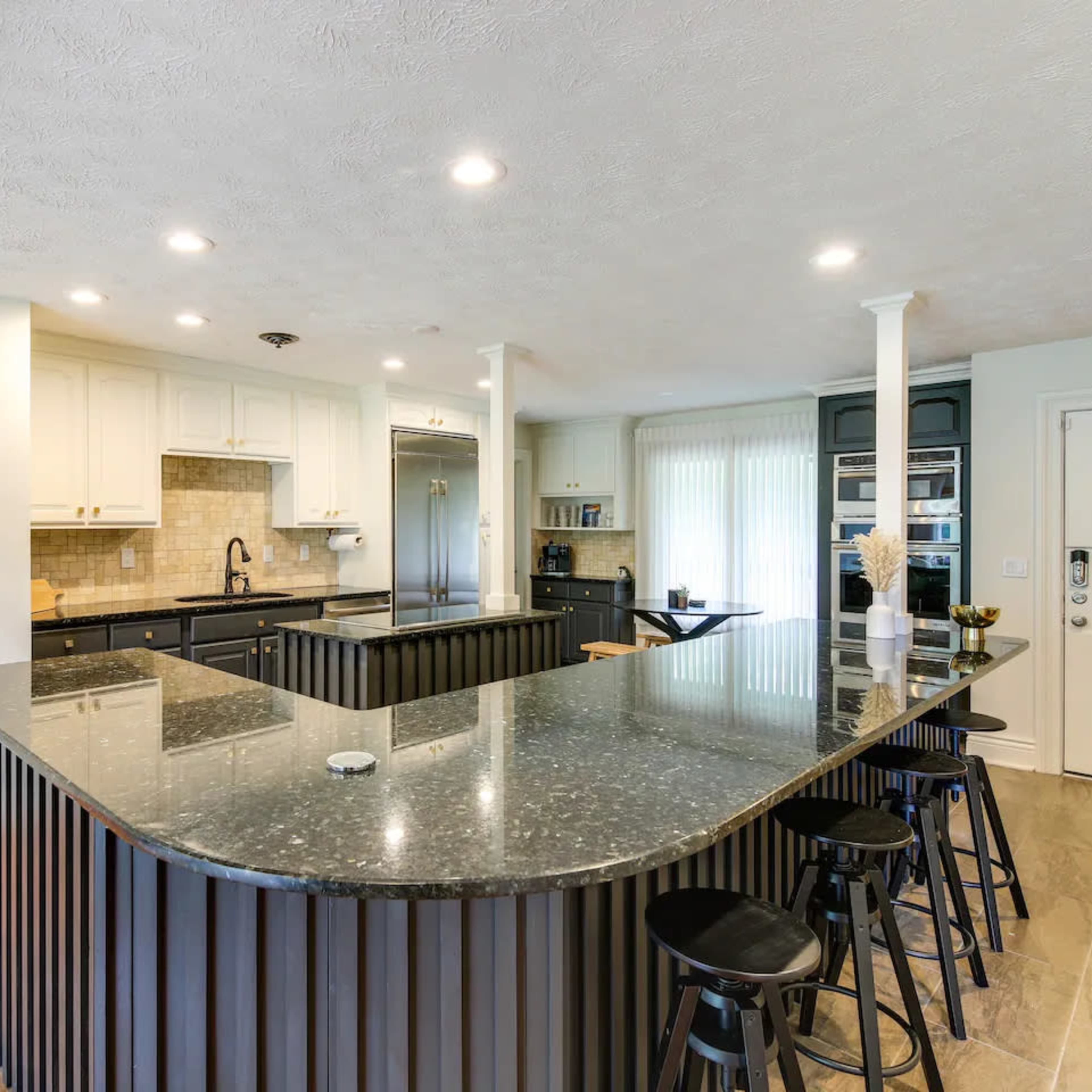 A modern kitchen with a large, dark granite island, black stools, white cabinetry, and a view of a dining space through an open doorway.