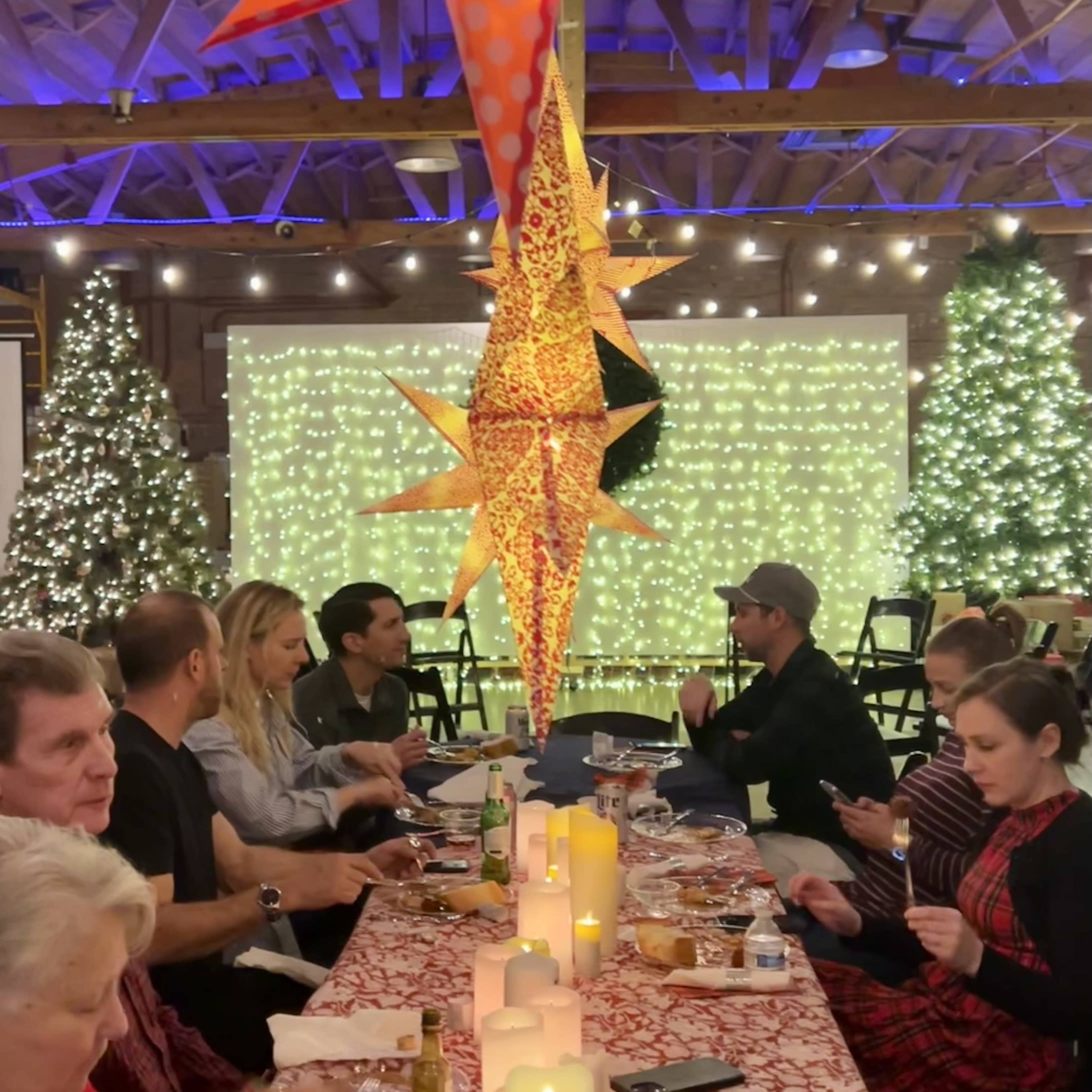 A spacious event venue with wooden beams, decorated with string lights and a festive backdrop, featuring tables arranged for a gathering.