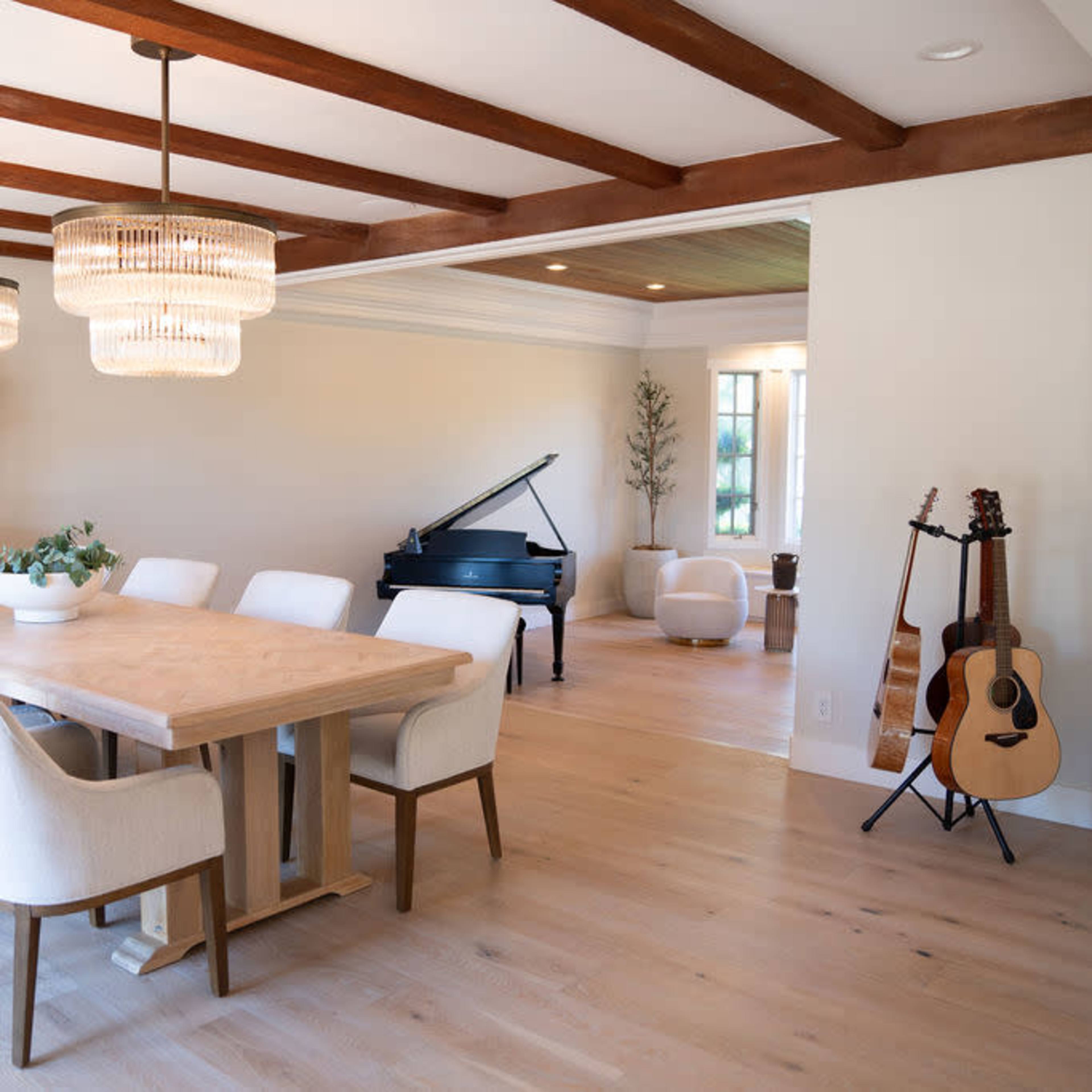 A modern dining area with a large wooden table surrounded by white chairs, a grand piano in the corner, and warm pendant lights overhead.
