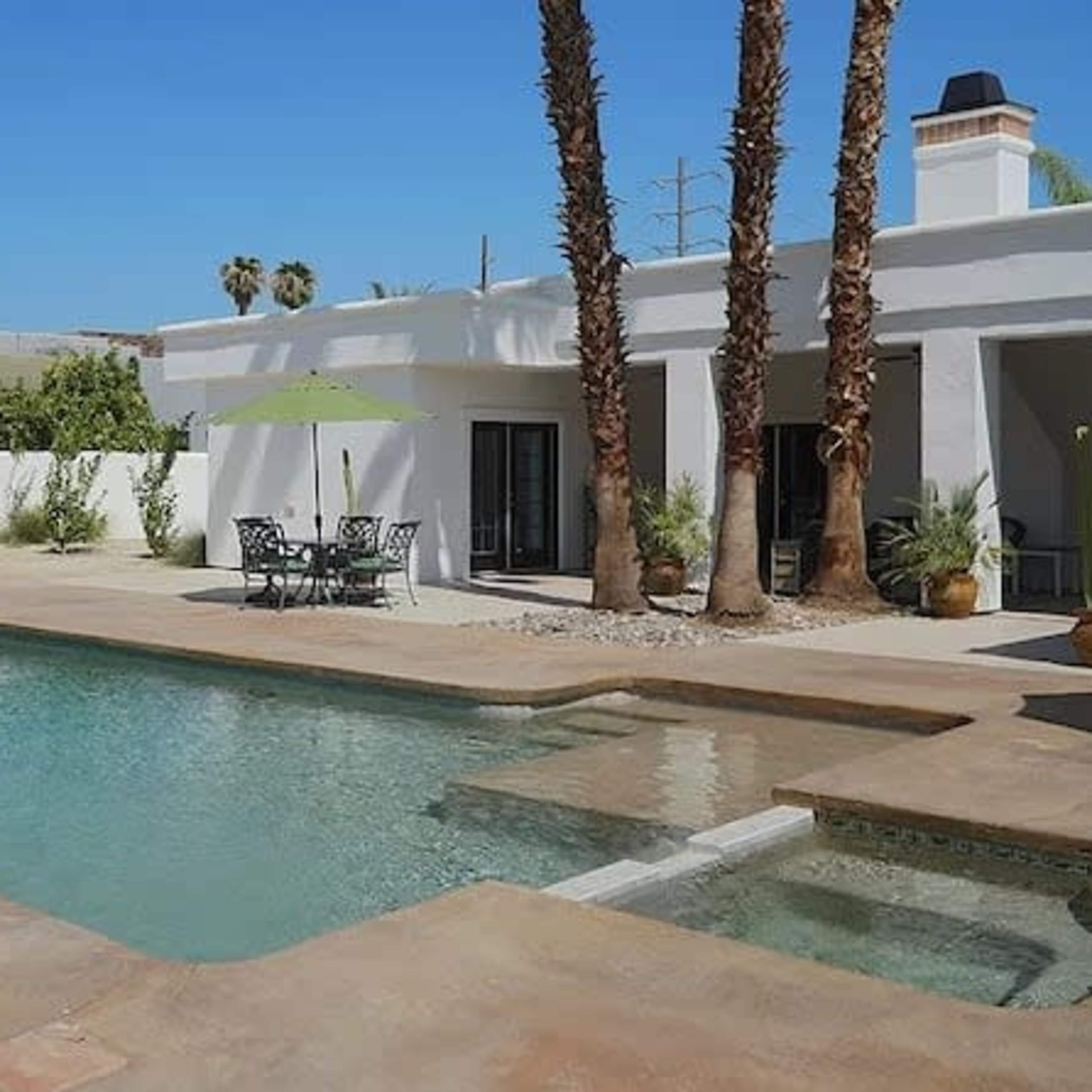 A swimming pool surrounded by lounge chairs and a table set under green umbrellas near a white building.