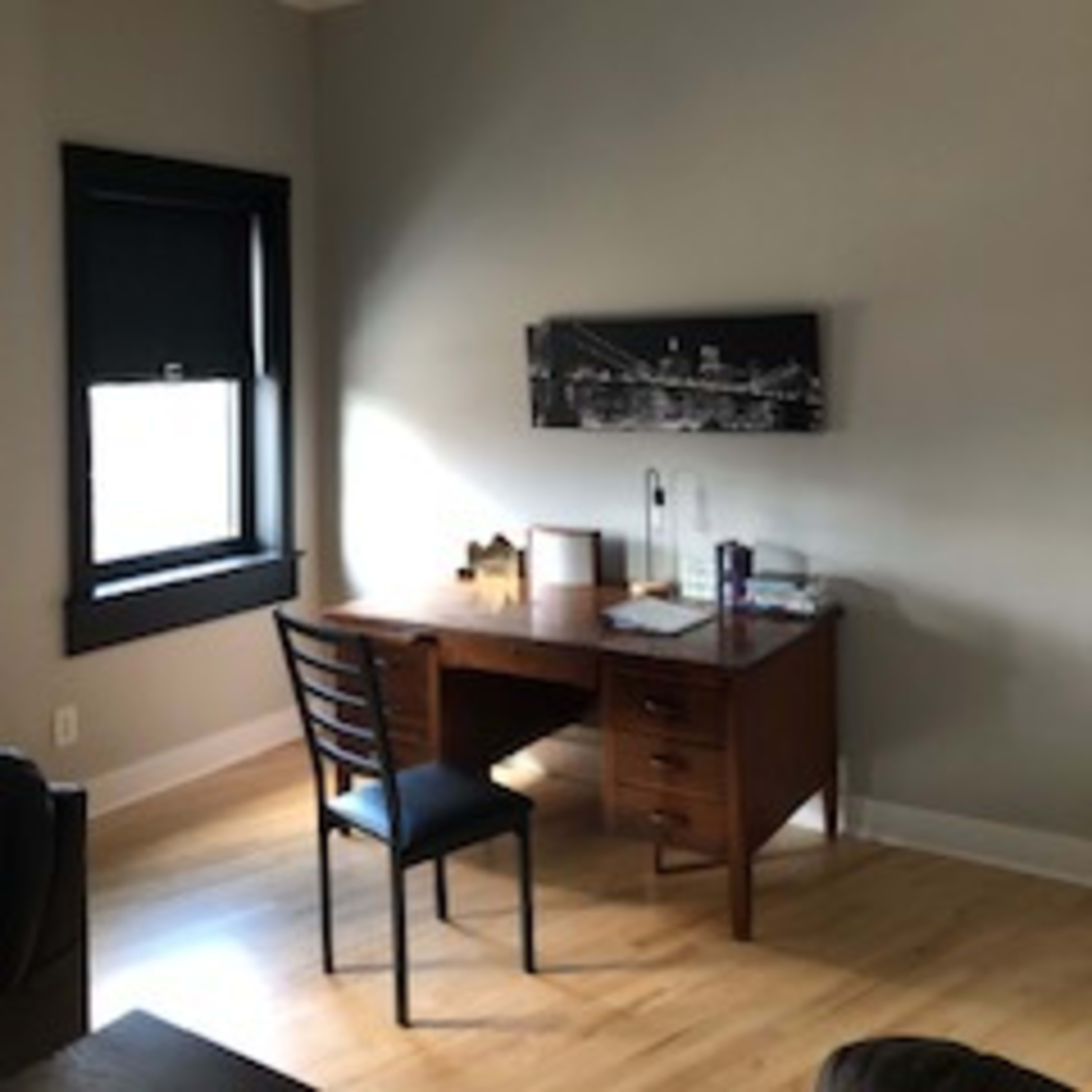 A simple home office with a wooden desk, a black chair, and a mounted black-and-white cityscape photo on the wall.
