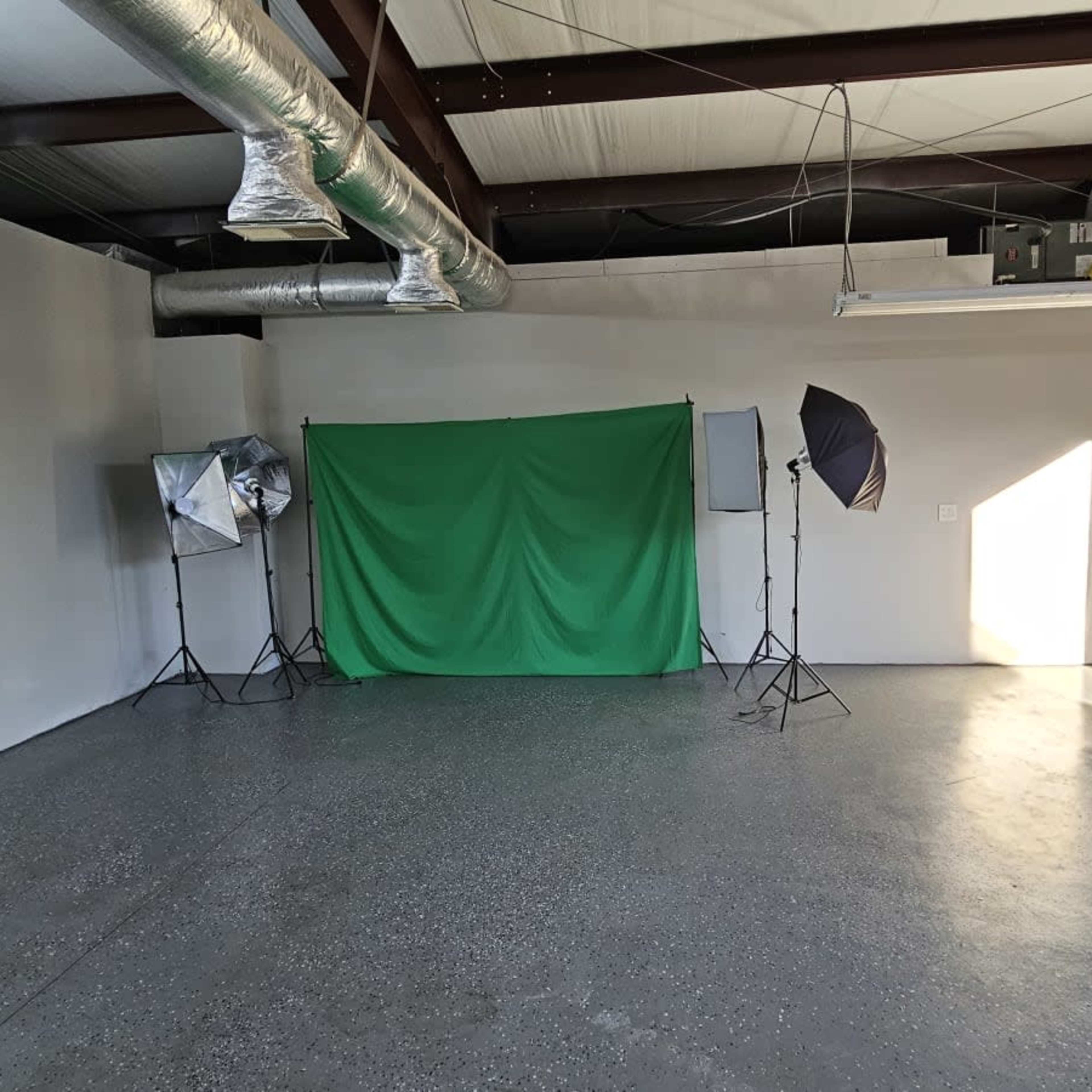 An empty studio space with a green backdrop, two softbox lights, and a window letting in natural light.