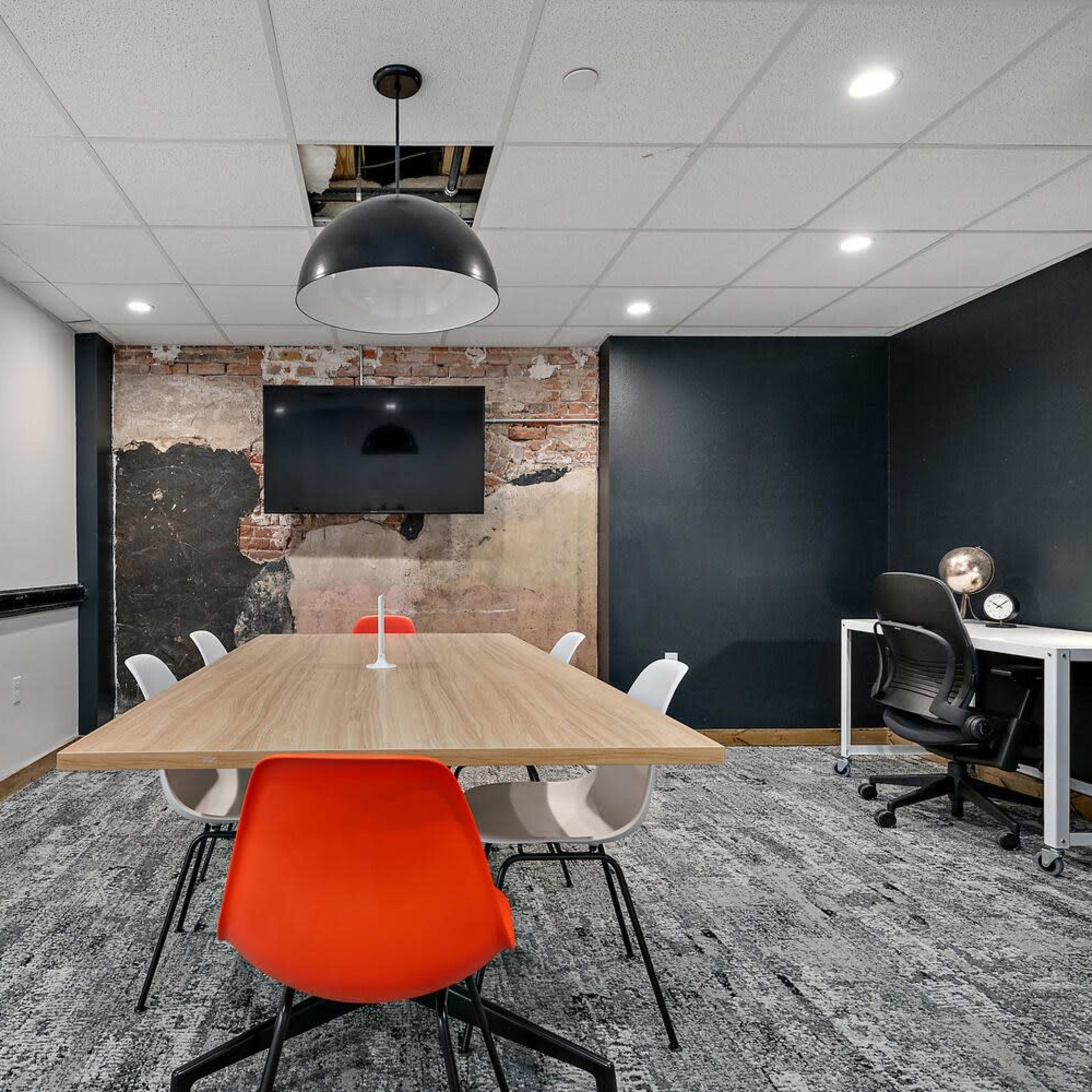 A modern meeting room features a large wooden table surrounded by white and orange chairs, with a black wall displaying a television and a section of exposed brick.