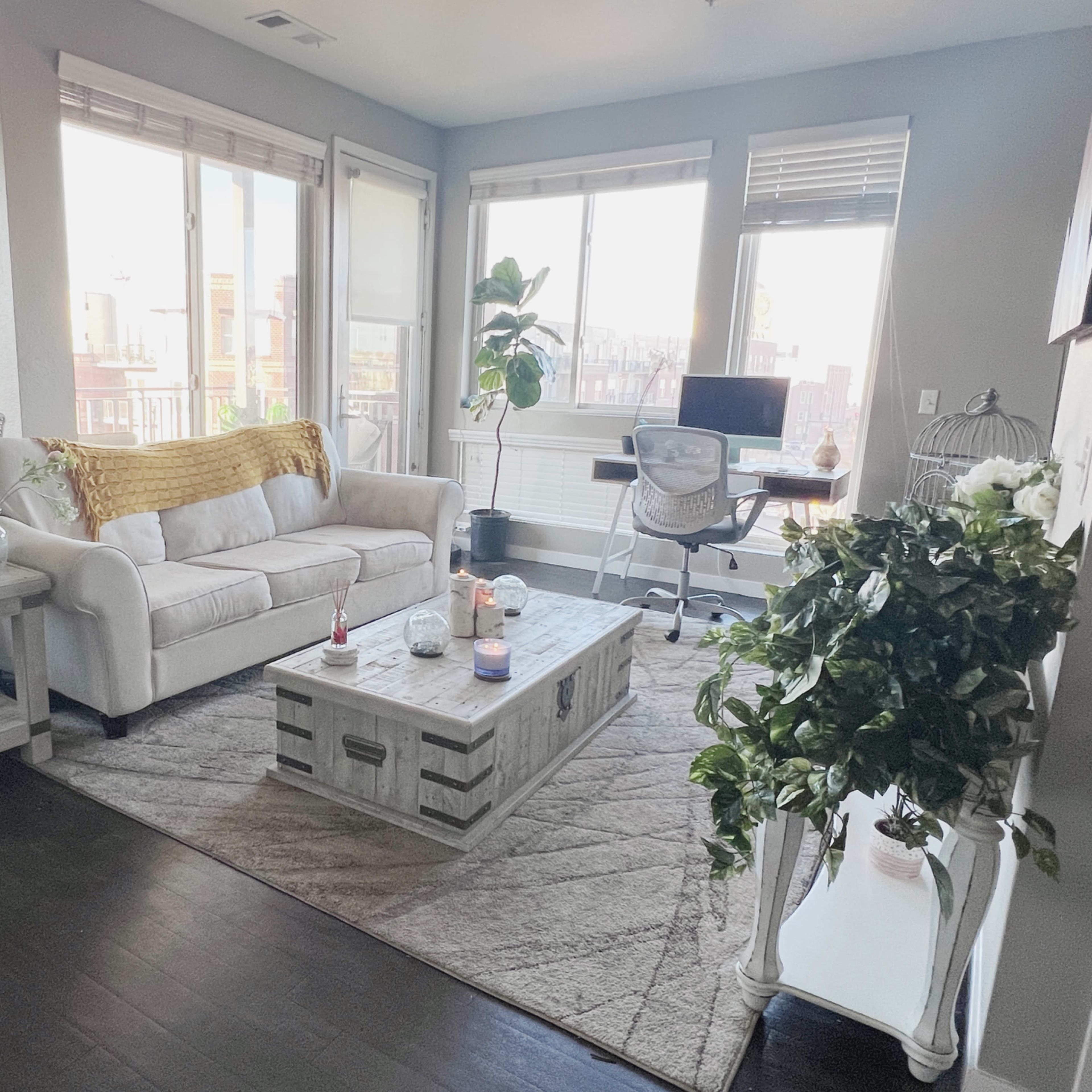 A bright living room with a white couch, a wooden coffee table, potted plants, and a desk with a chair near large windows.