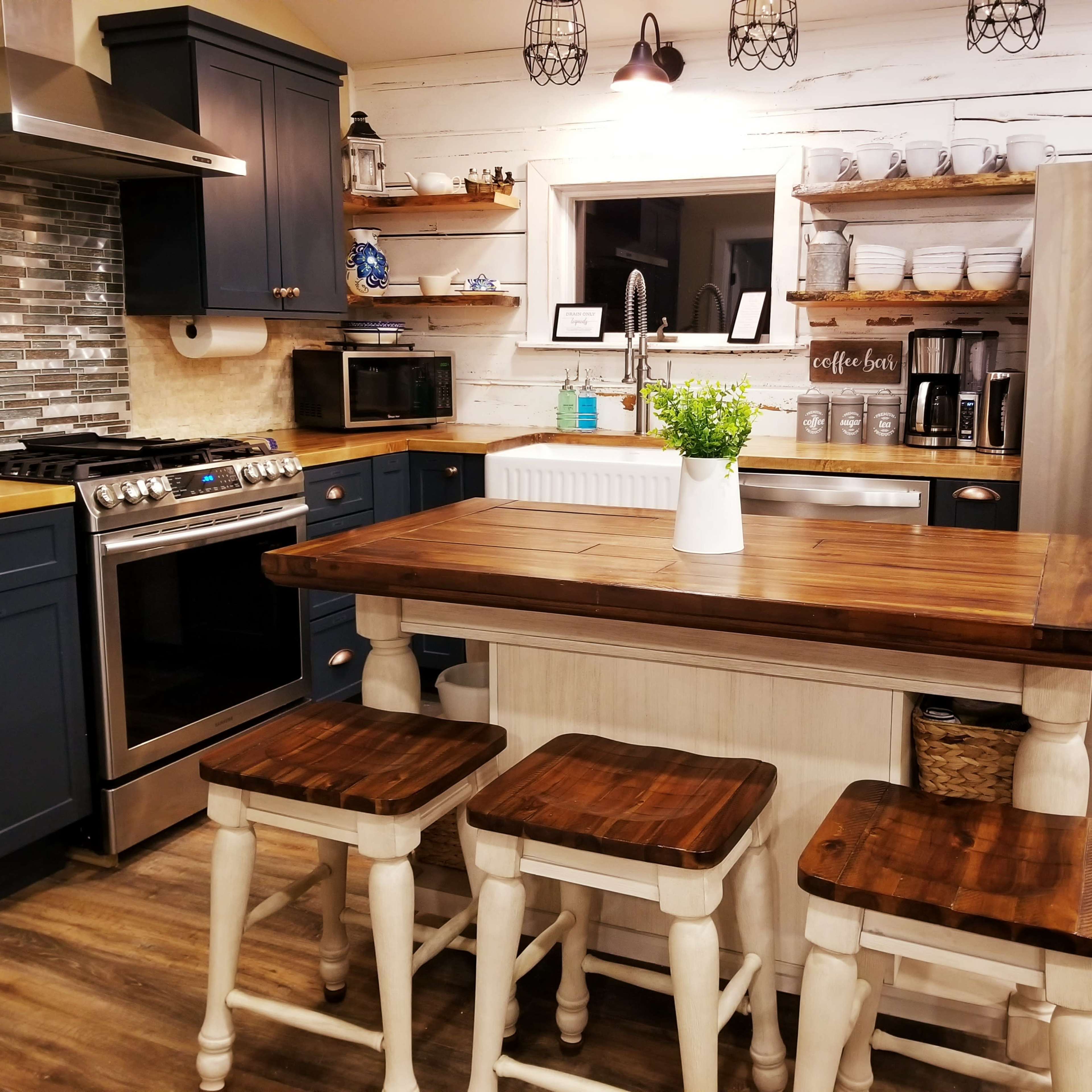 A modern kitchen featuring a central wooden island with three stools, stainless steel appliances, and dark blue cabinetry against a light, rustic wall.