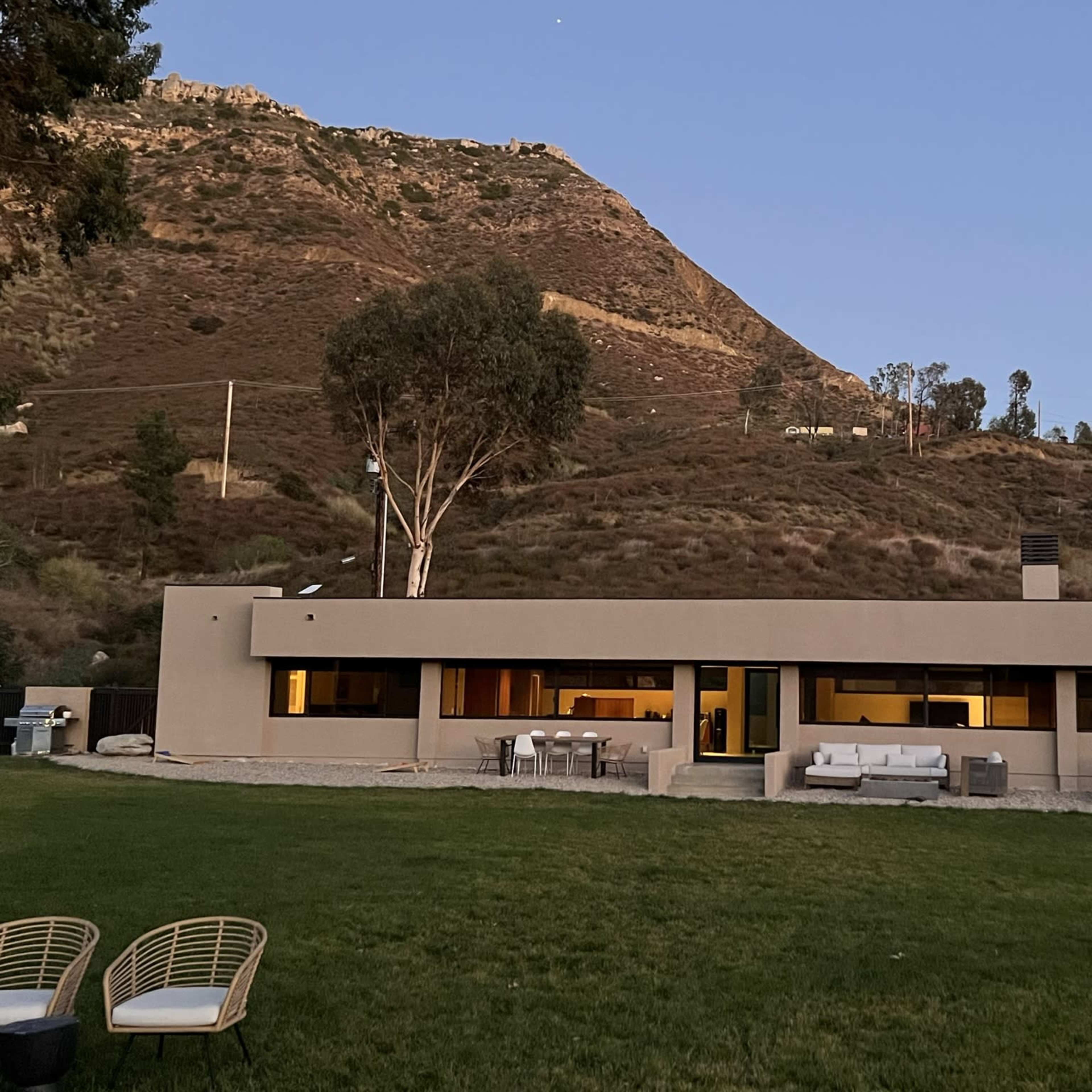 A modern single-story house situated on a grassy area with a hillside in the background at dusk.