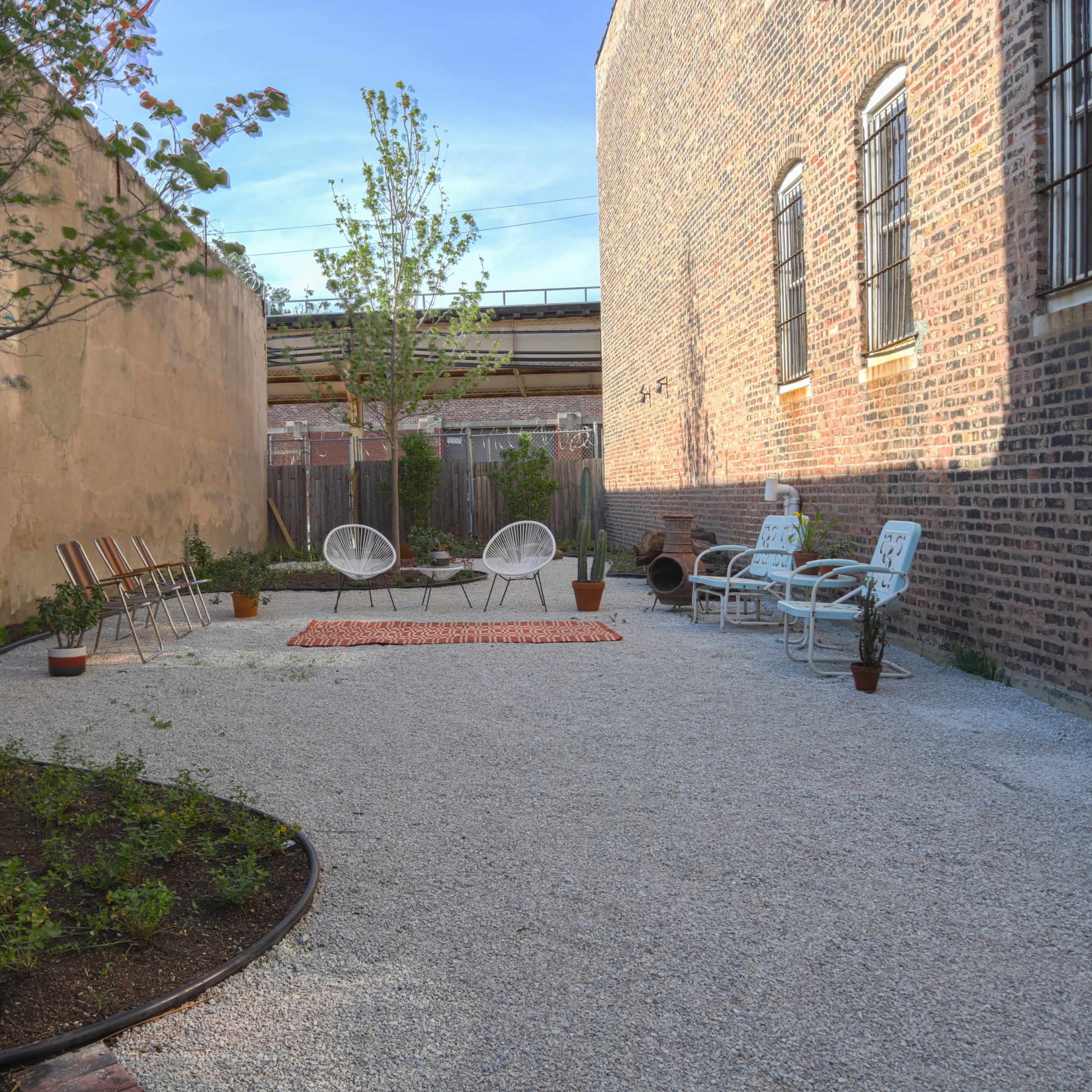 A narrow outdoor space with gravel flooring, featuring seating arrangements, potted plants, and a small area rug surrounded by brick walls.