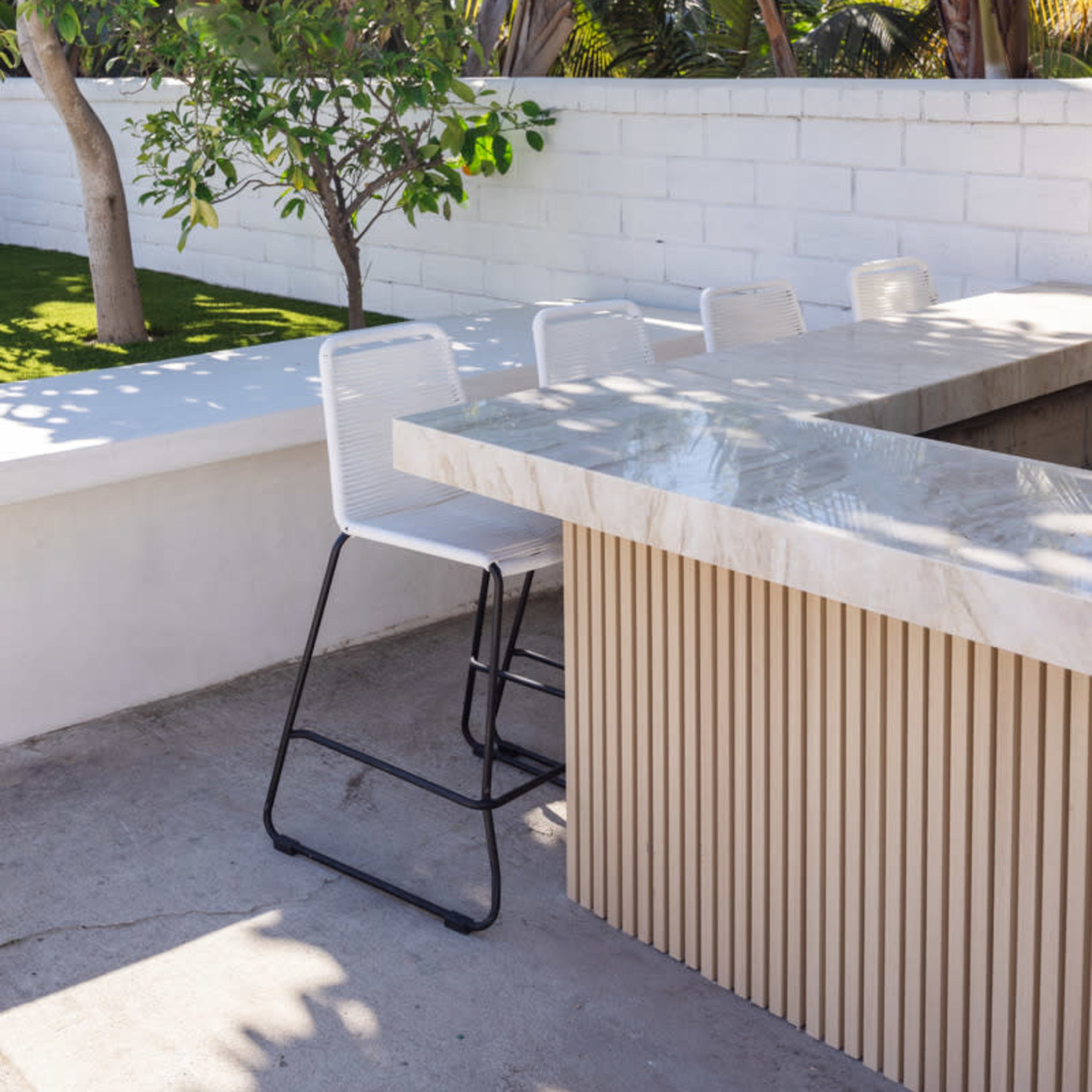 A modern outdoor bar area with a marble countertop and white bar stools, surrounded by lush greenery and a white wall.