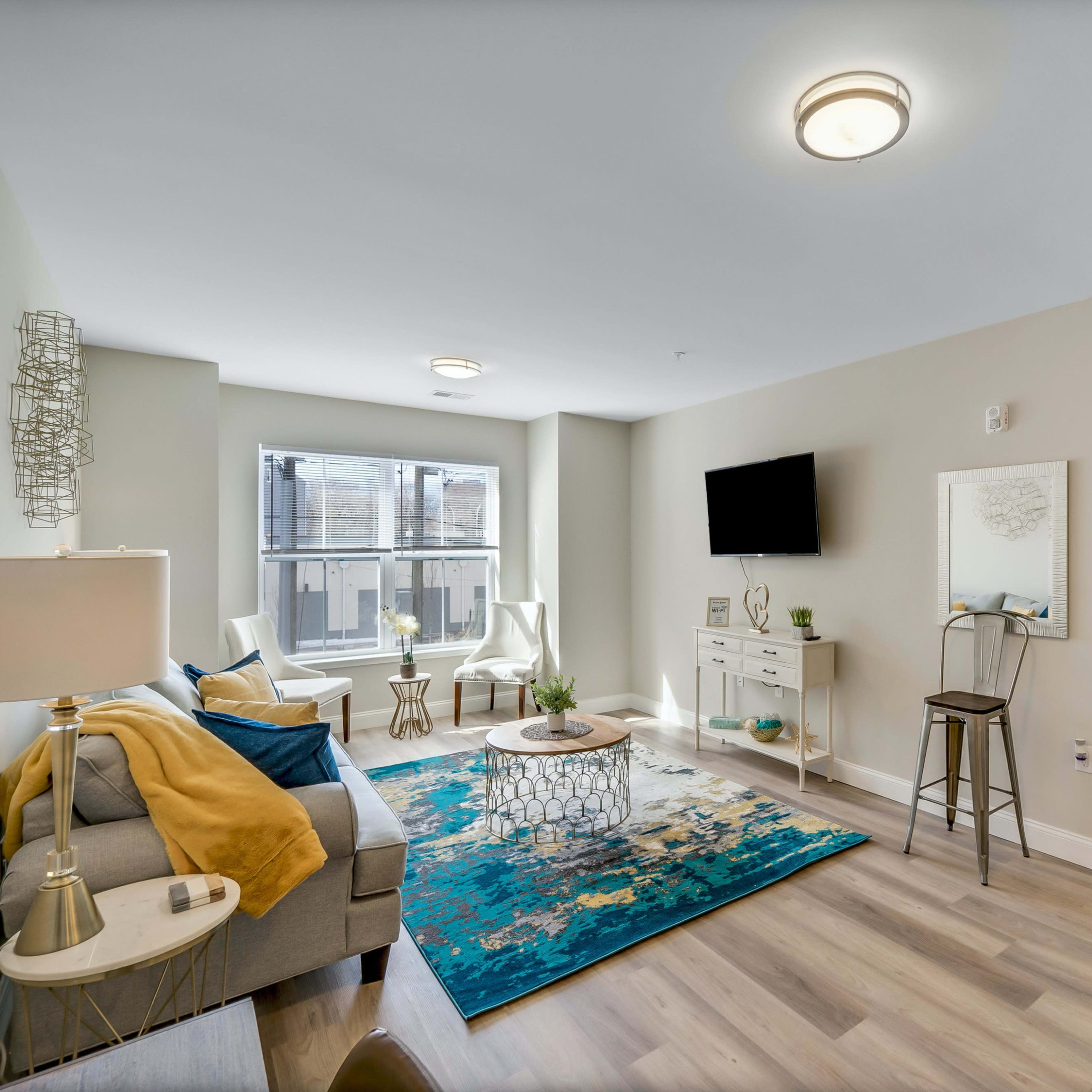 A modern living room with a light-colored sofa, a coffee table on a patterned rug, and a wall-mounted TV, along with a small dining area visible in the background.