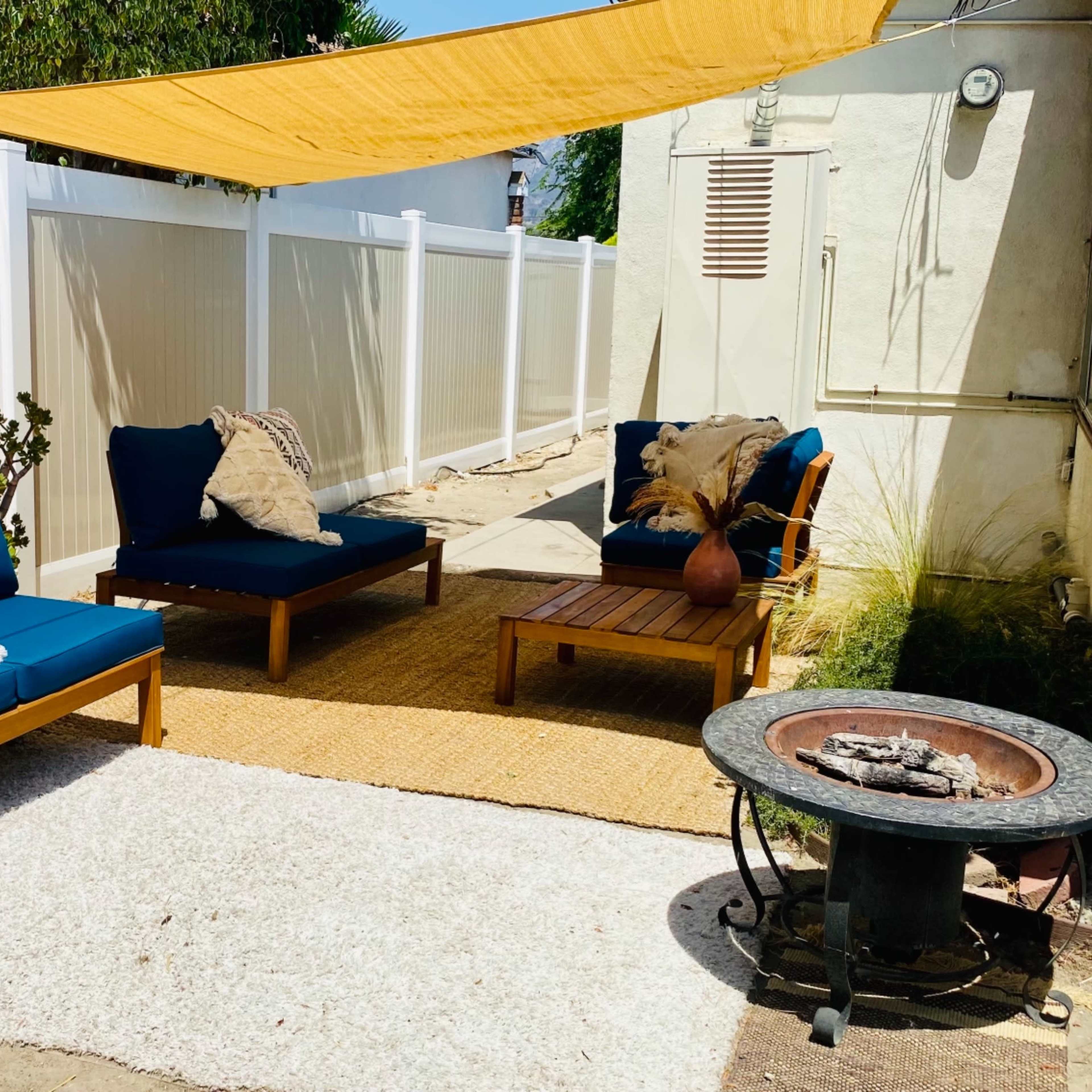 A cozy outdoor seating area with two blue lounge chairs, a small fire pit, and a rug under a yellow canopy near a white fence.