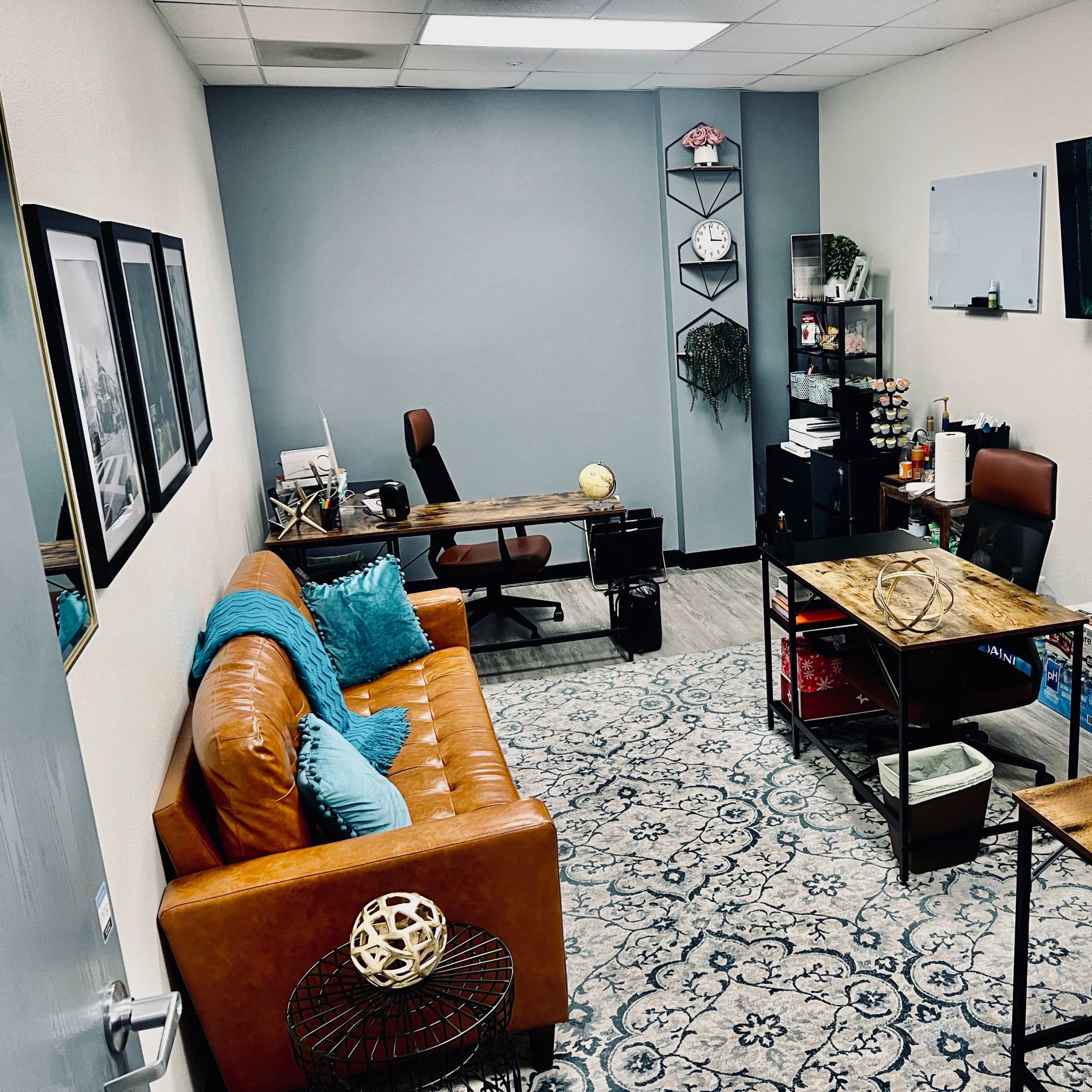 A modern office space with a brown leather sofa, two desks, a television on the wall, and decorative elements like a globe and plants.