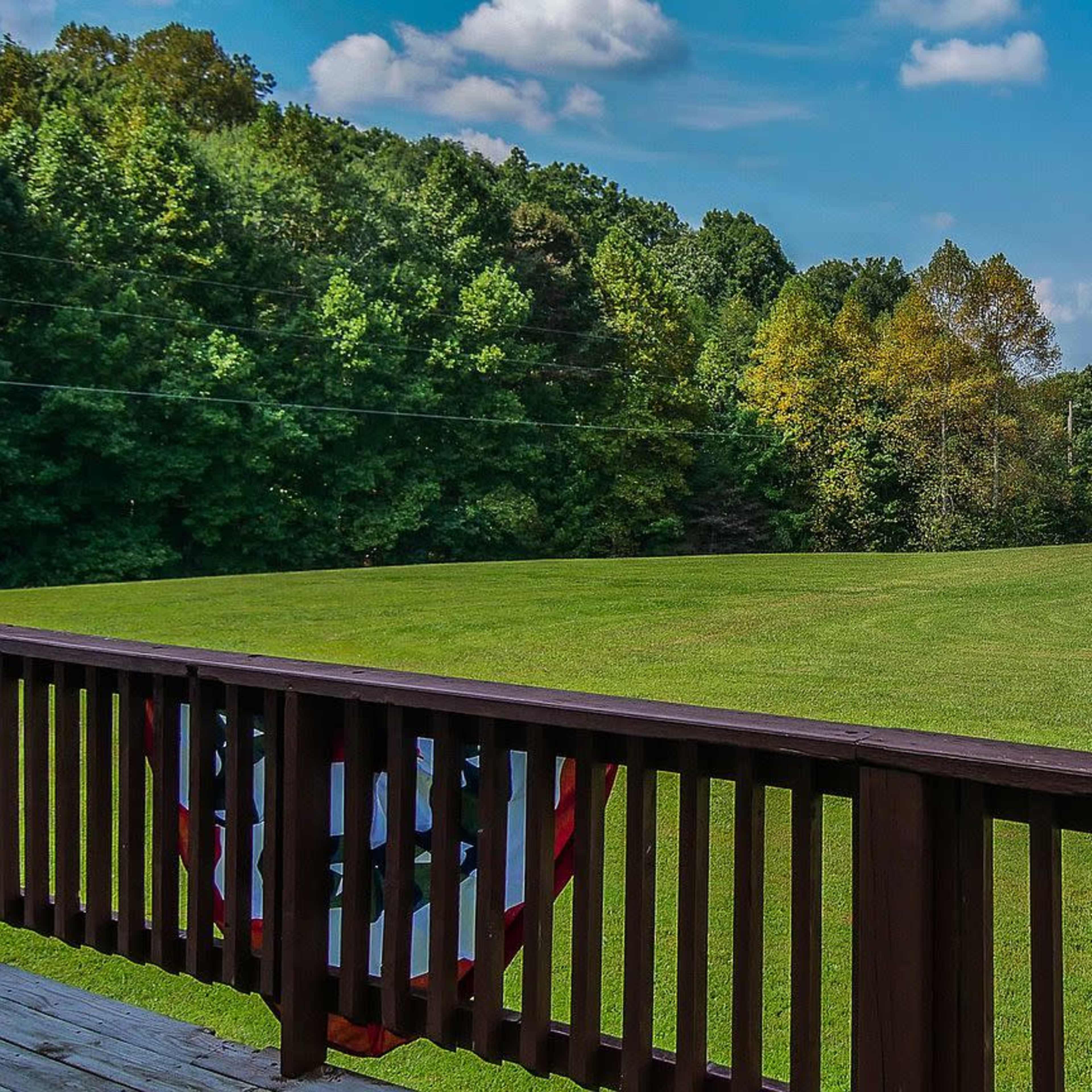 A wooden deck overlooks a large, grassy field bordered by trees under a blue sky with scattered clouds.