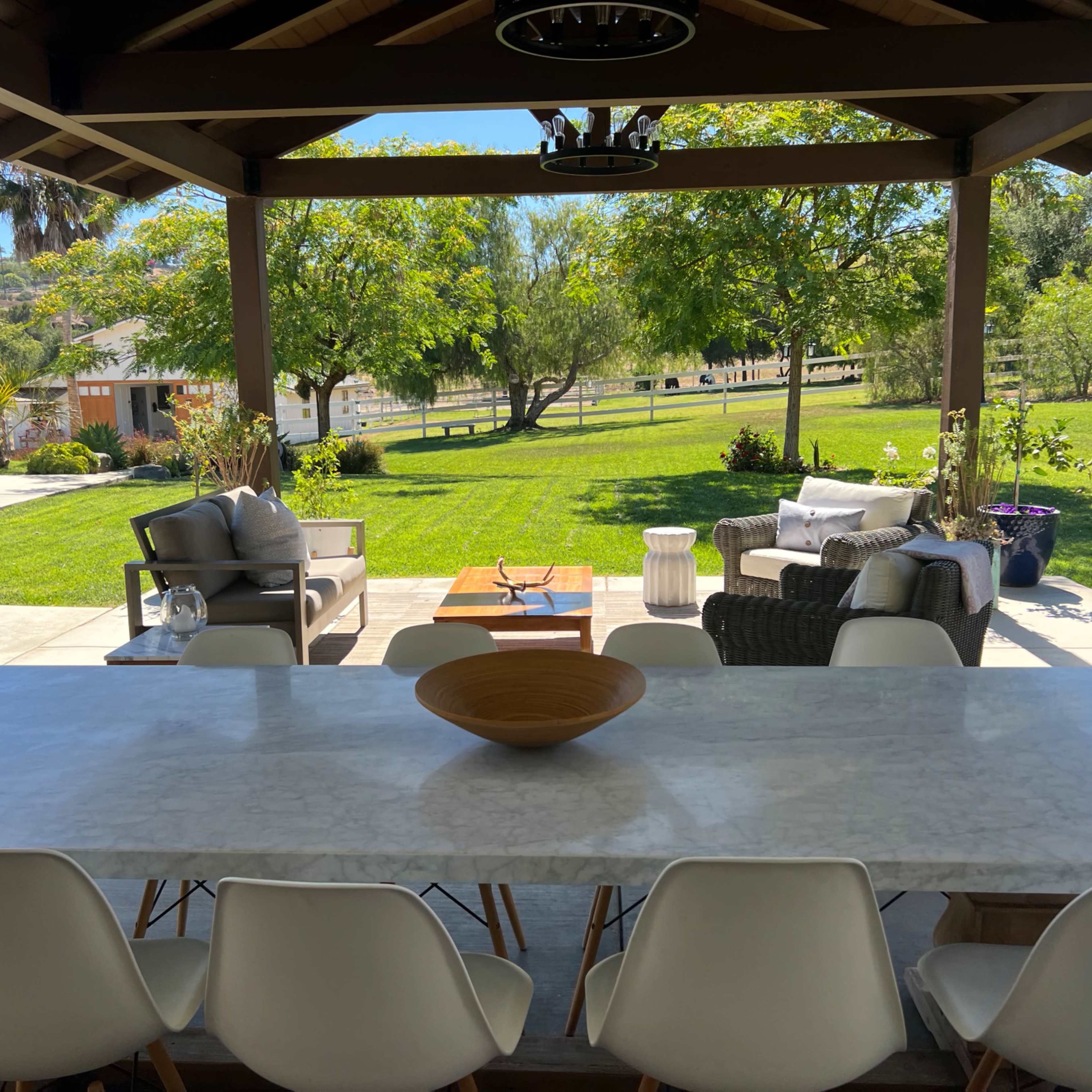 A spacious outdoor dining area features a long marble table and comfortable seating, overlooking a well-maintained lawn and trees.