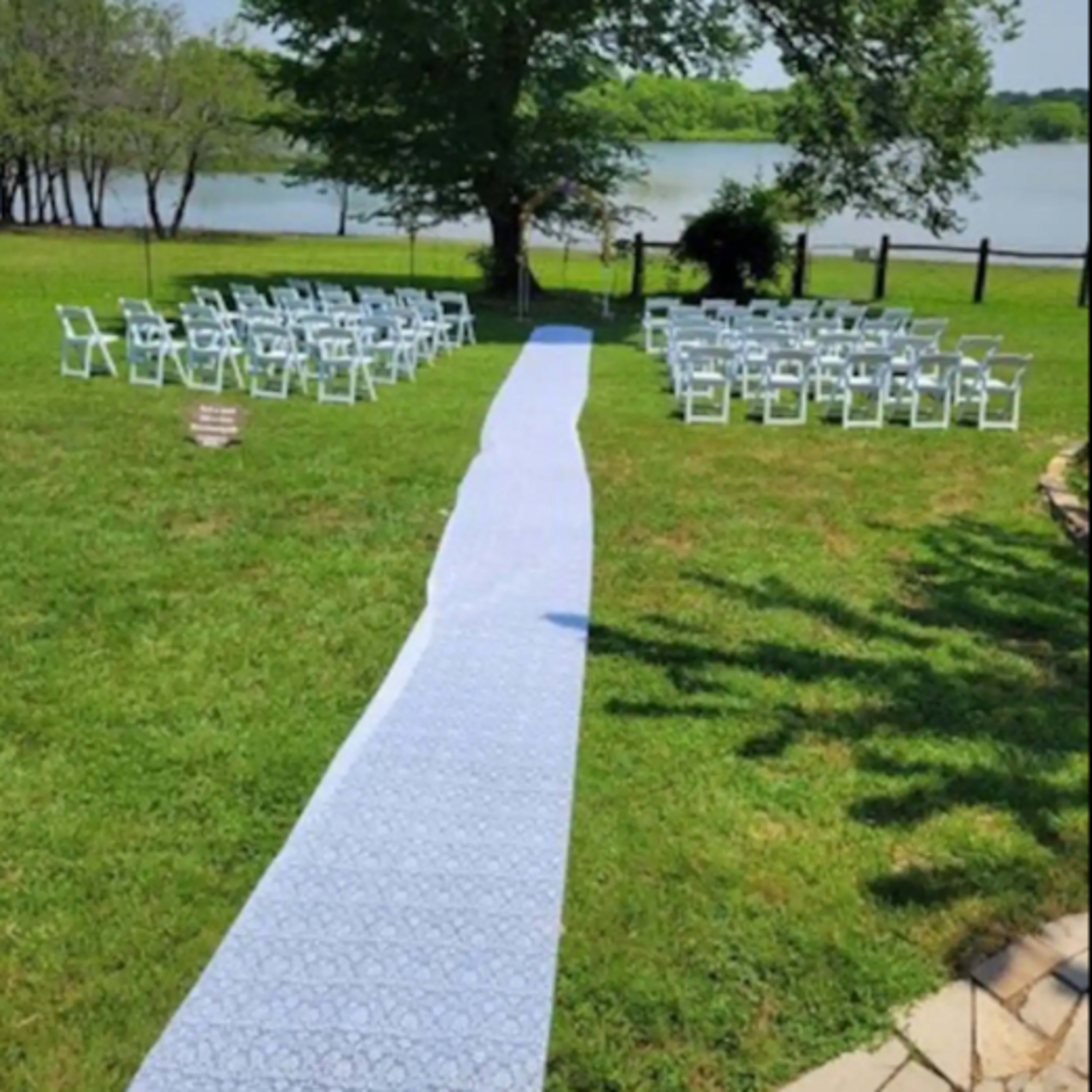 A wedding setup featuring a blue patterned aisle runner leading to rows of white chairs arranged on a grassy area by a lake.