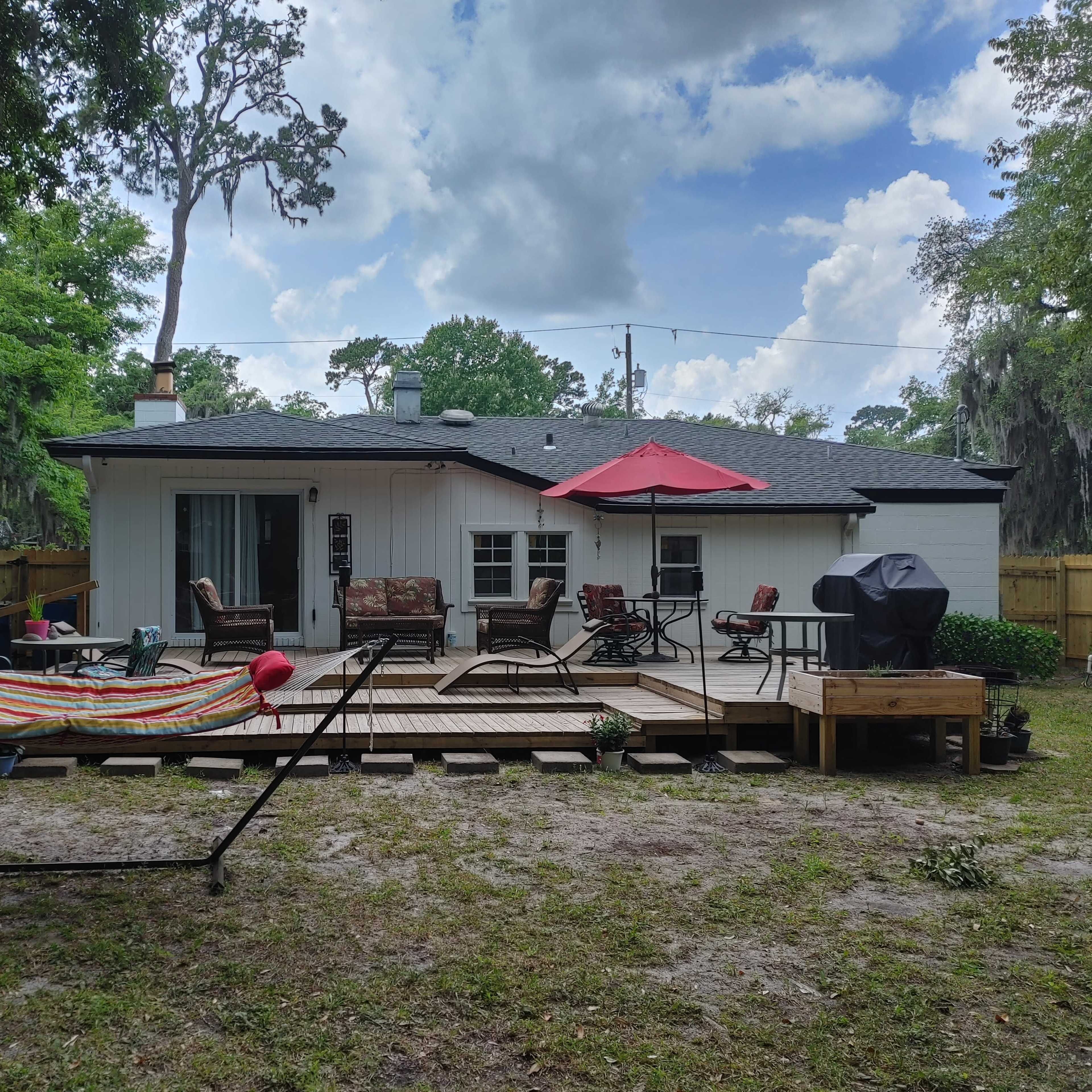 A backyard with a wooden deck, a hammock, seating area, and a barbecue grill, set against a backdrop of trees and a partly cloudy sky.