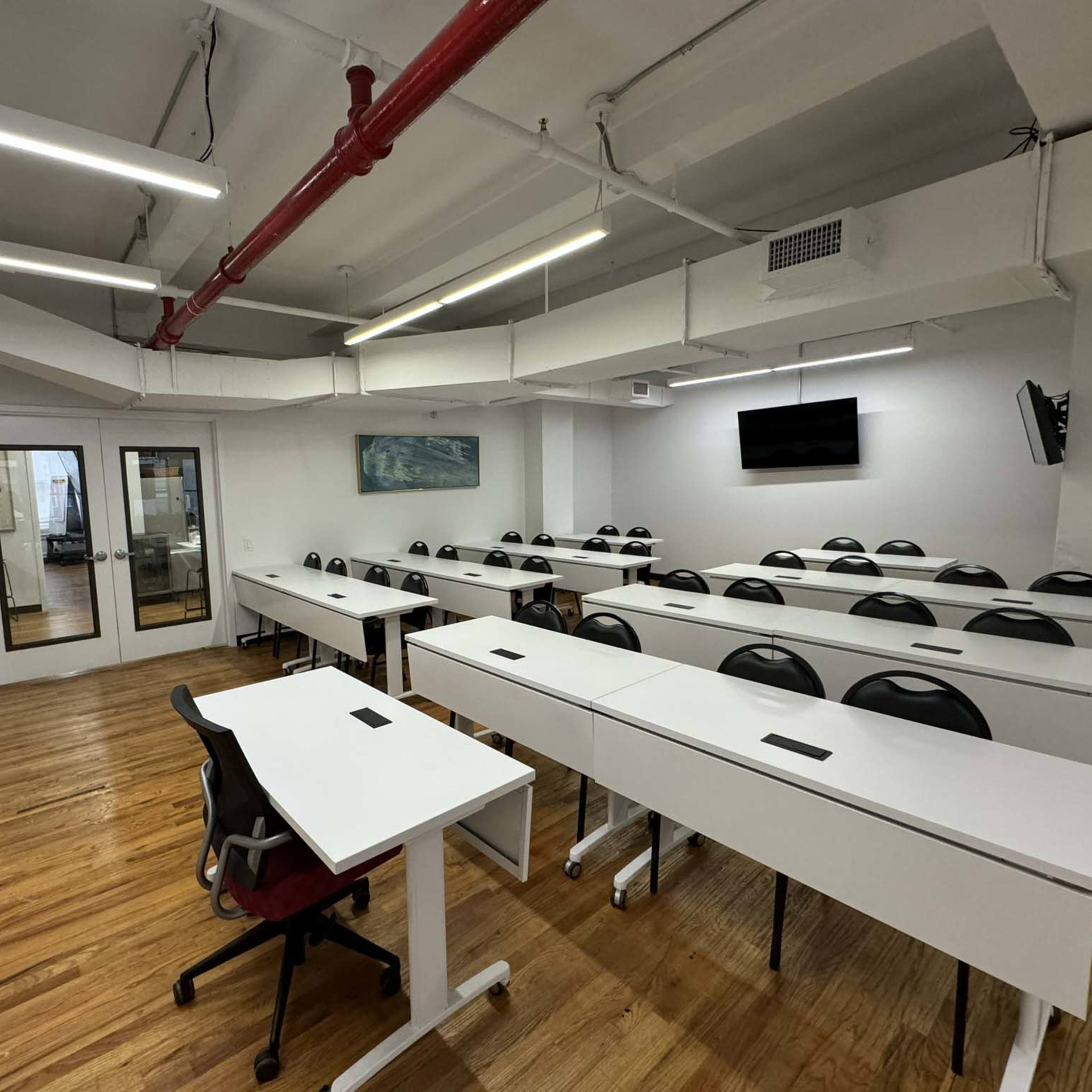 A bright classroom features multiple rows of desks and chairs, with two large screens on the walls and windows leading to an adjacent space.