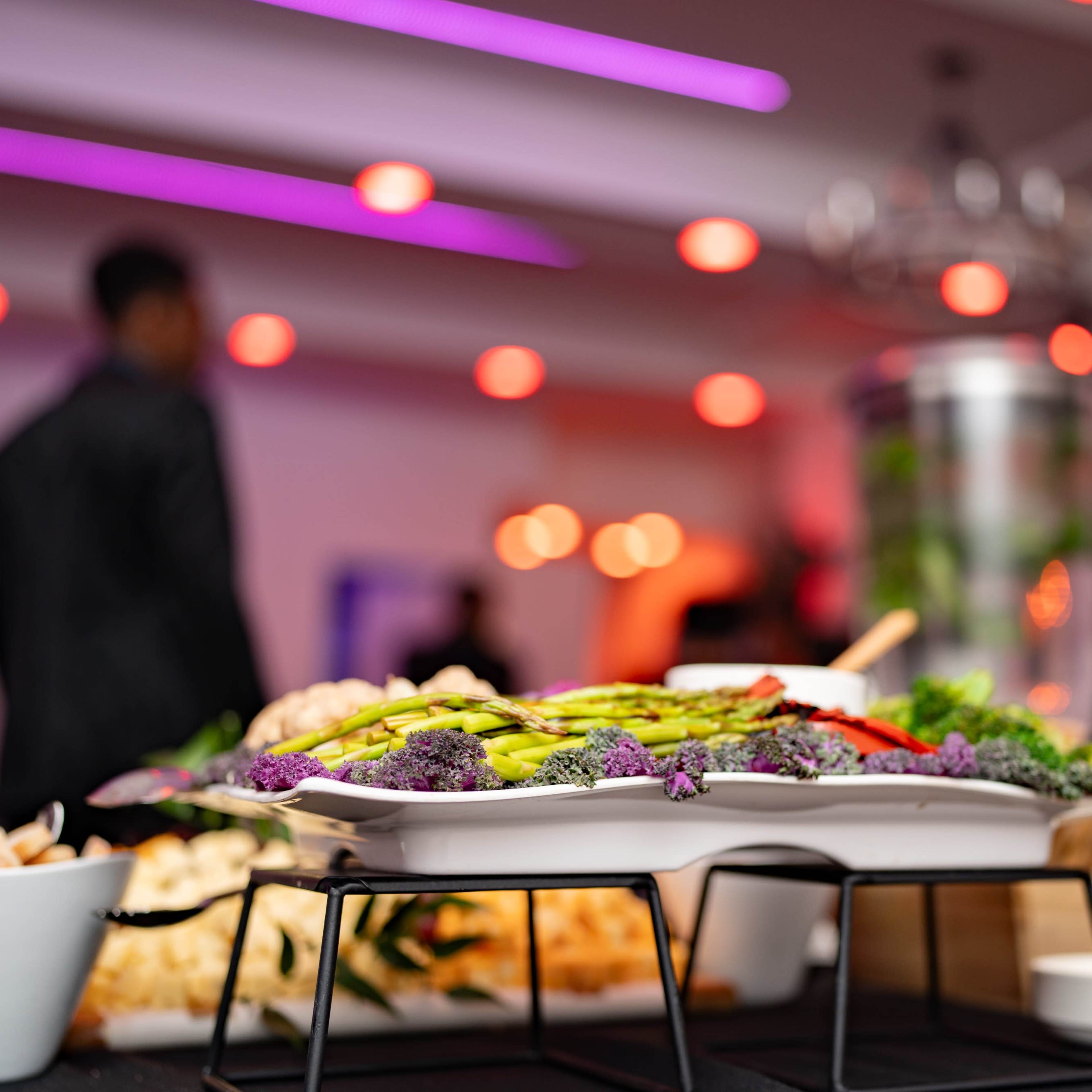 A beautifully arranged platter of vegetables and snacks is set on a table, with blurred figures and soft lighting in the background.