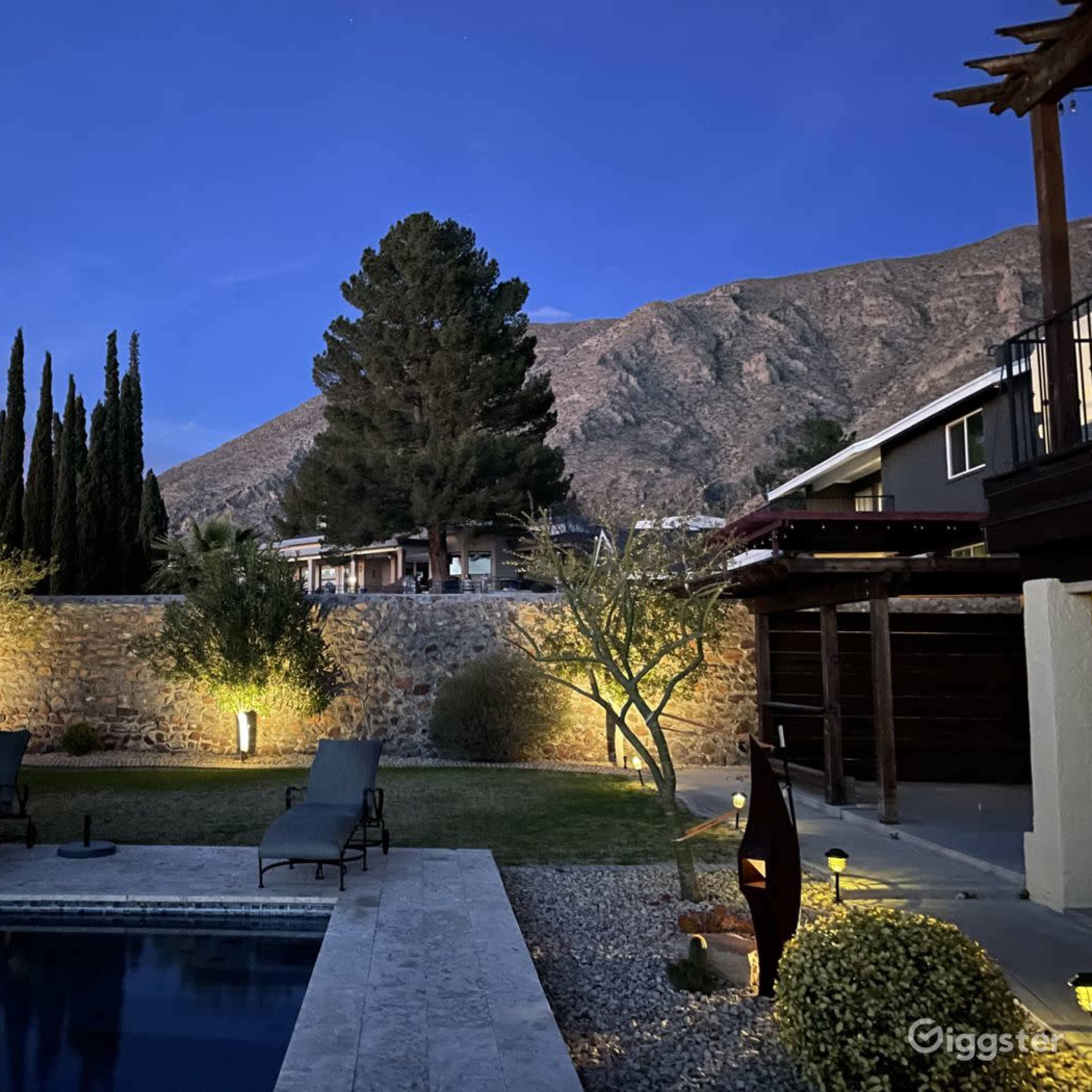 A serene backyard with a pool, lounge chairs, and illuminated trees, set against a backdrop of mountains at twilight.