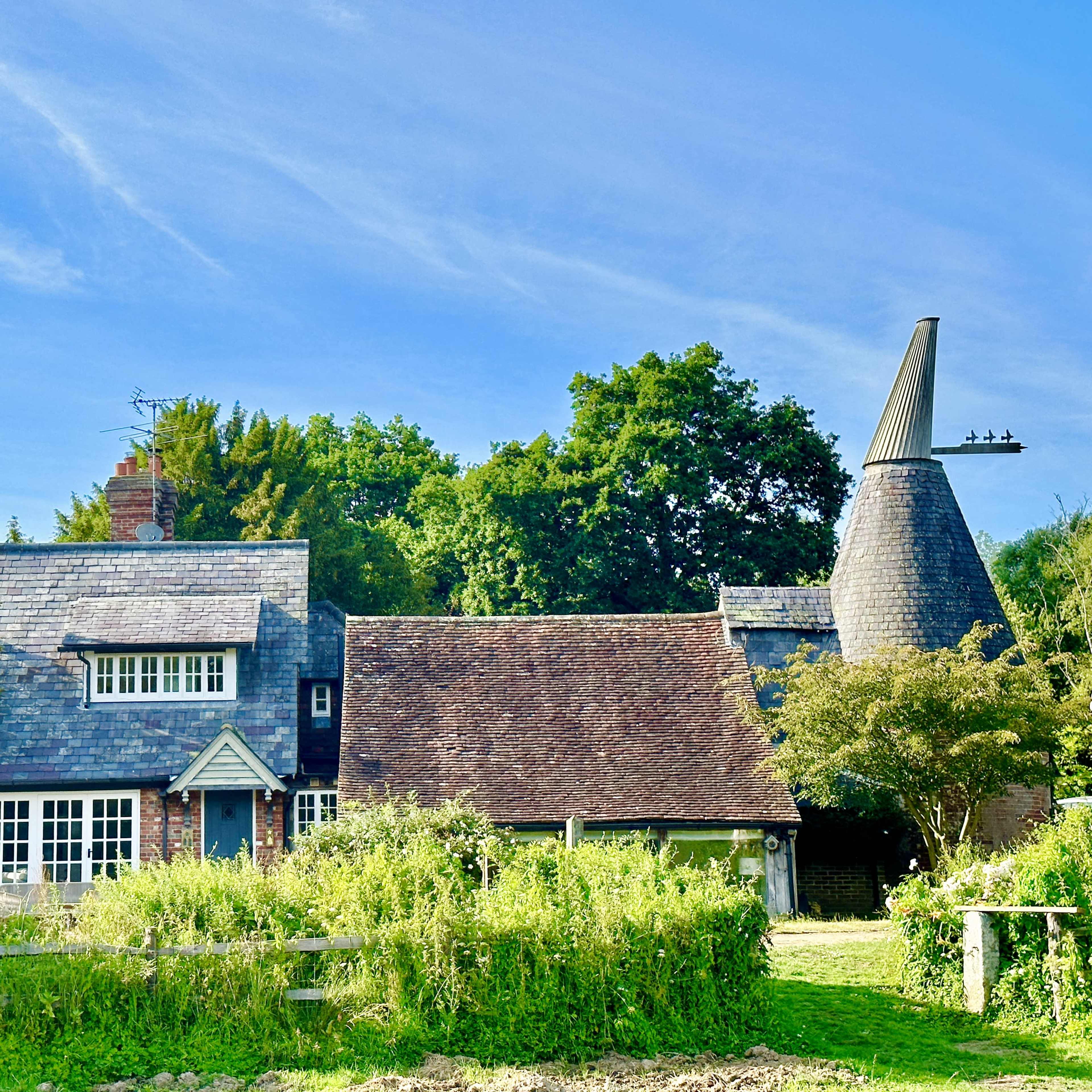 A quaint house with a conical roof structure surrounded by lush greenery and trees under a clear blue sky.