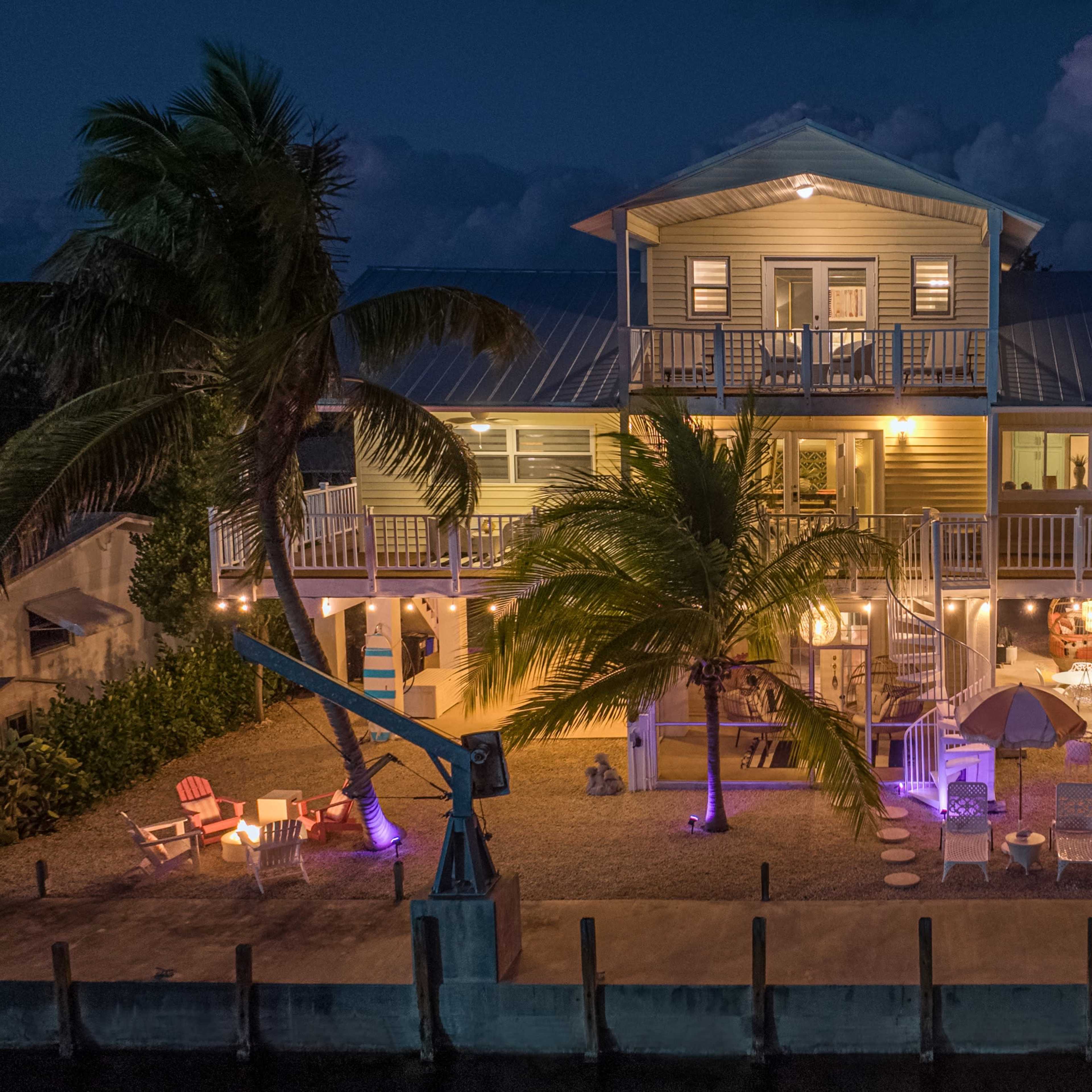A two-story waterfront house with a balcony and outdoor seating is illuminated by lights at night, surrounded by palm trees.