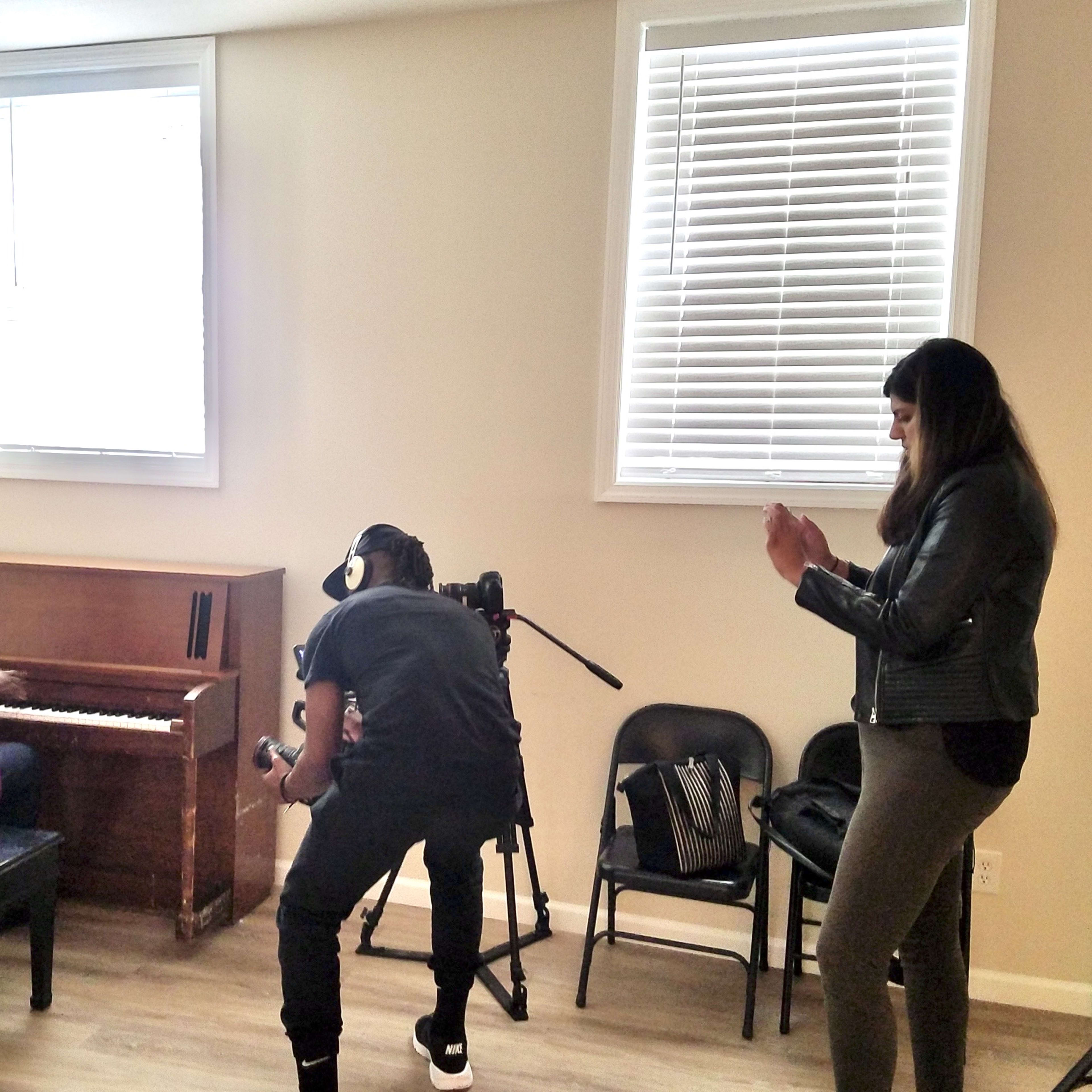 A person in a purple suit plays a piano while being filmed by a cameraman, and a woman stands nearby clapping.