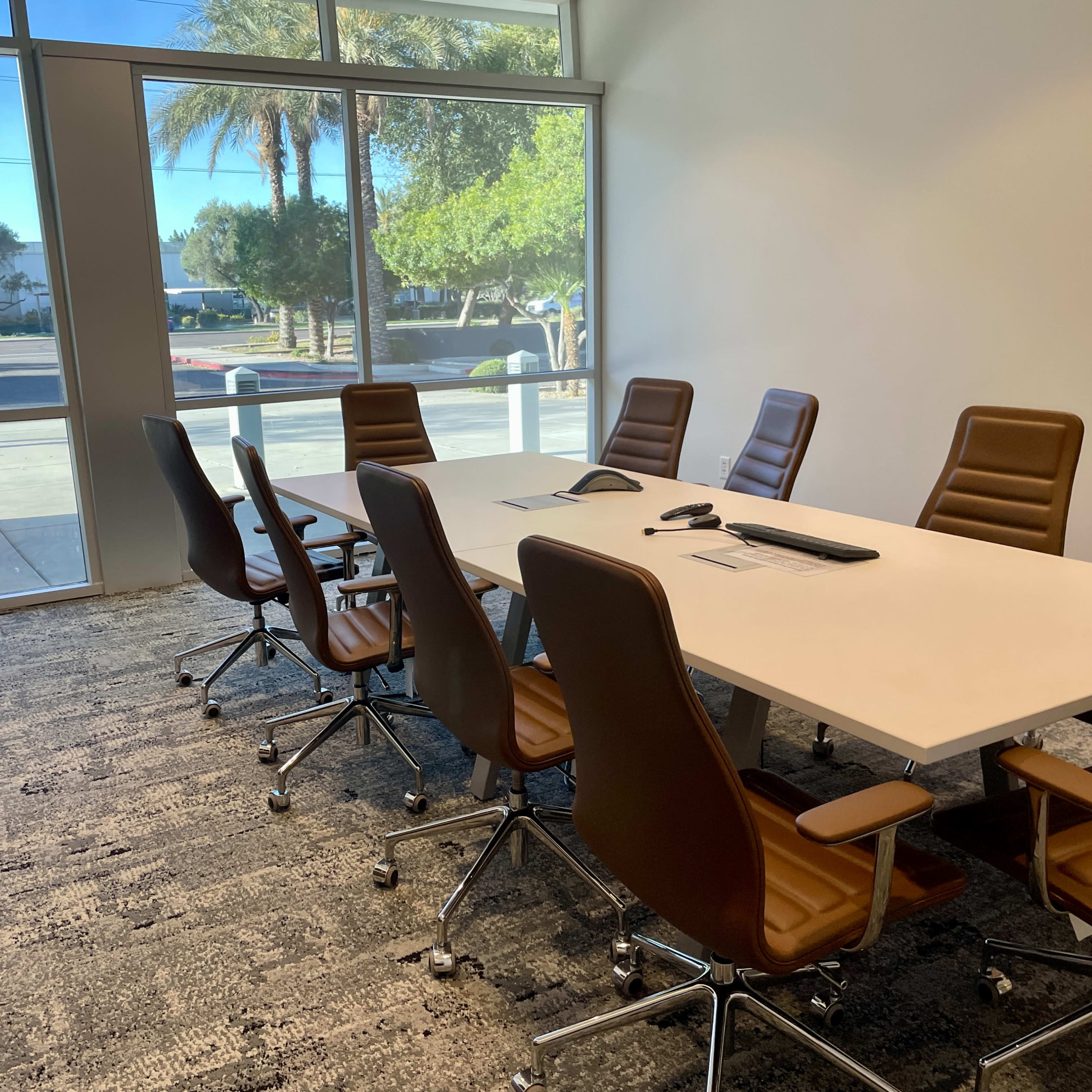 A conference room features a large white table surrounded by brown leather chairs, with large windows allowing natural light to enter.
