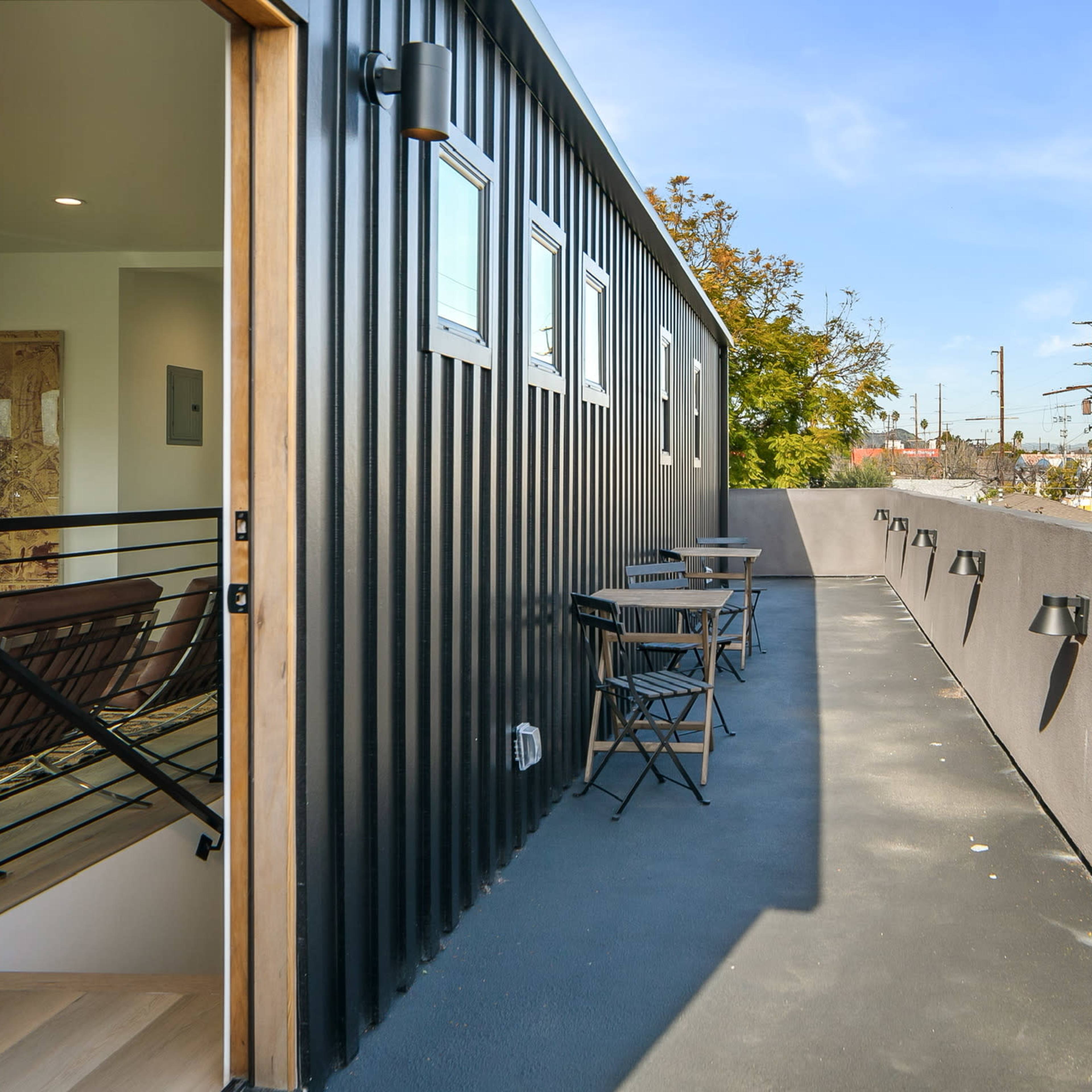 A narrow outdoor balcony with metal siding and small tables, overlooking an industrial area with telephone poles and trees in the background.