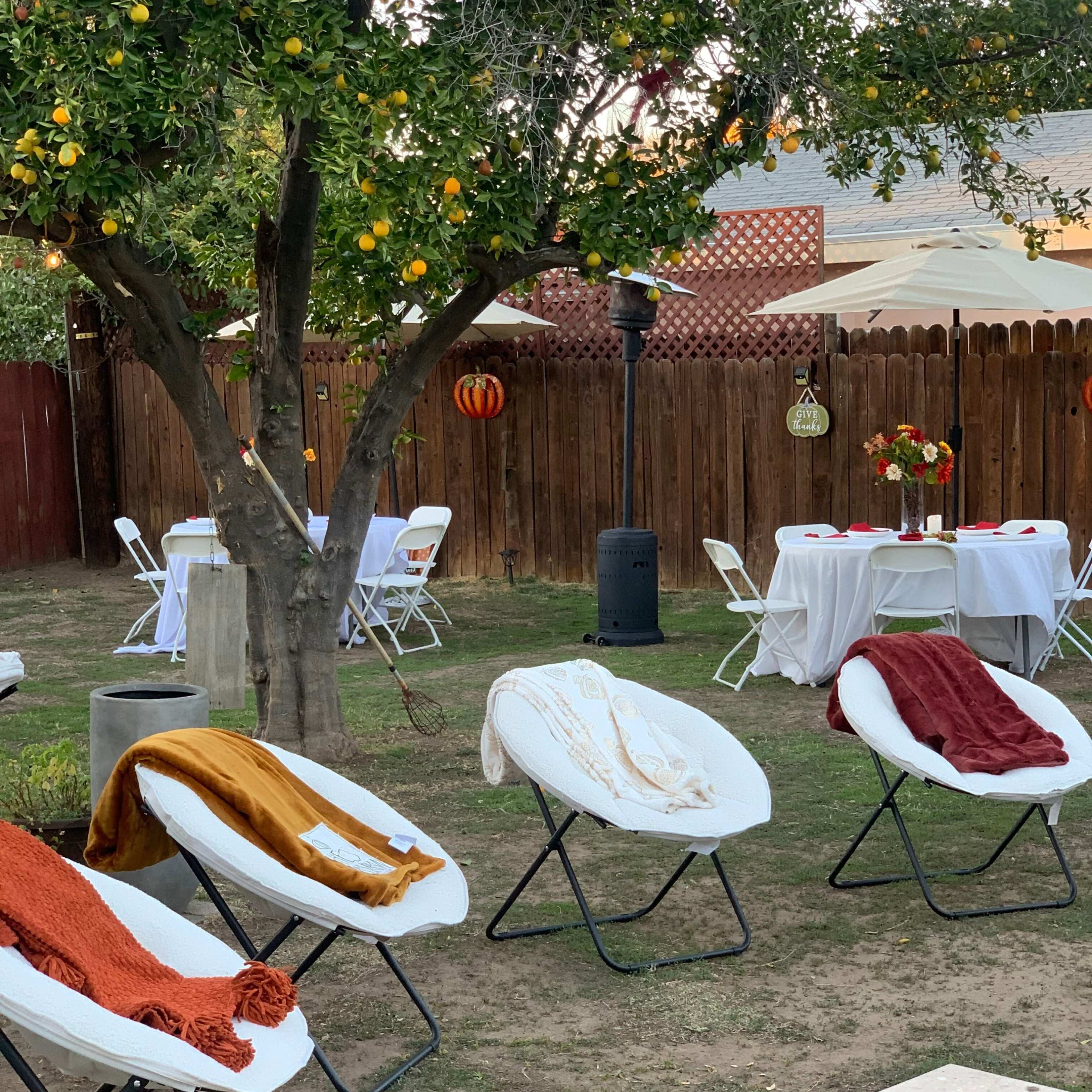 A backyard setting with several white lounge chairs arranged on grass, a table setup with white tablecloths and chairs under a tree with hanging decorations, and a wooden fence in the background.