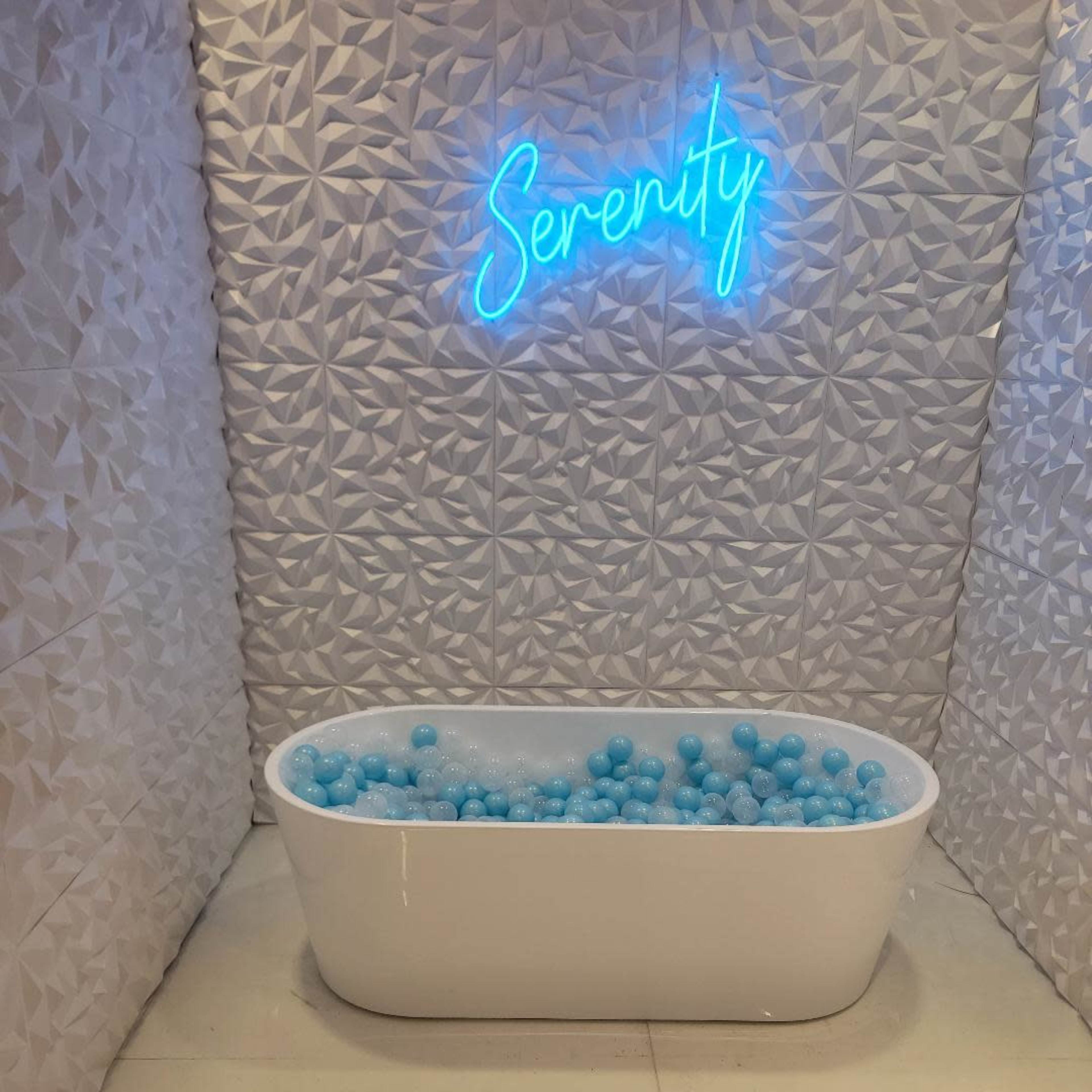 A white bathtub filled with blue balls sits beneath a neon sign reading "Serenity" against a textured white wall.