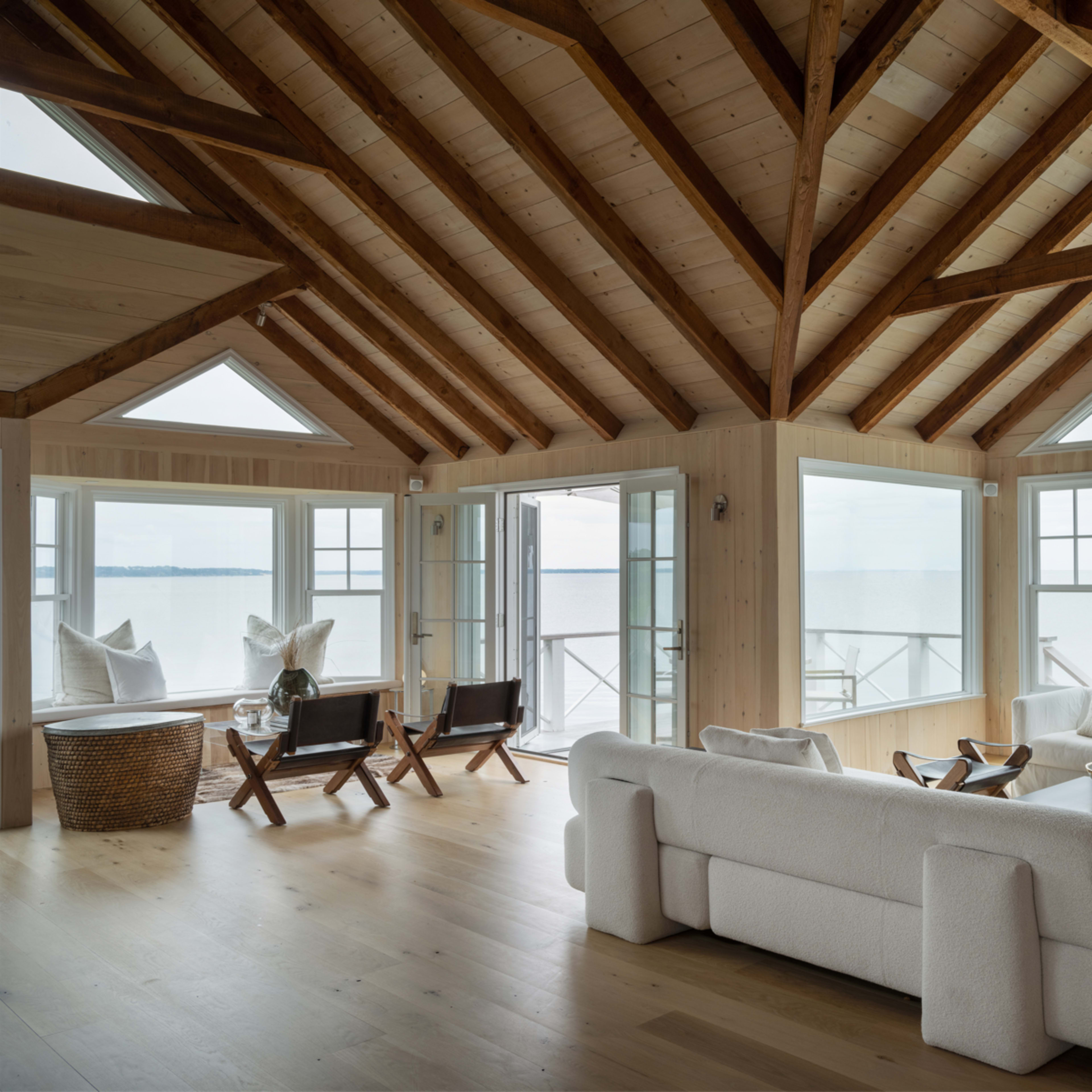 A spacious, light-filled living room with wooden beams, large windows, and a view of the water.