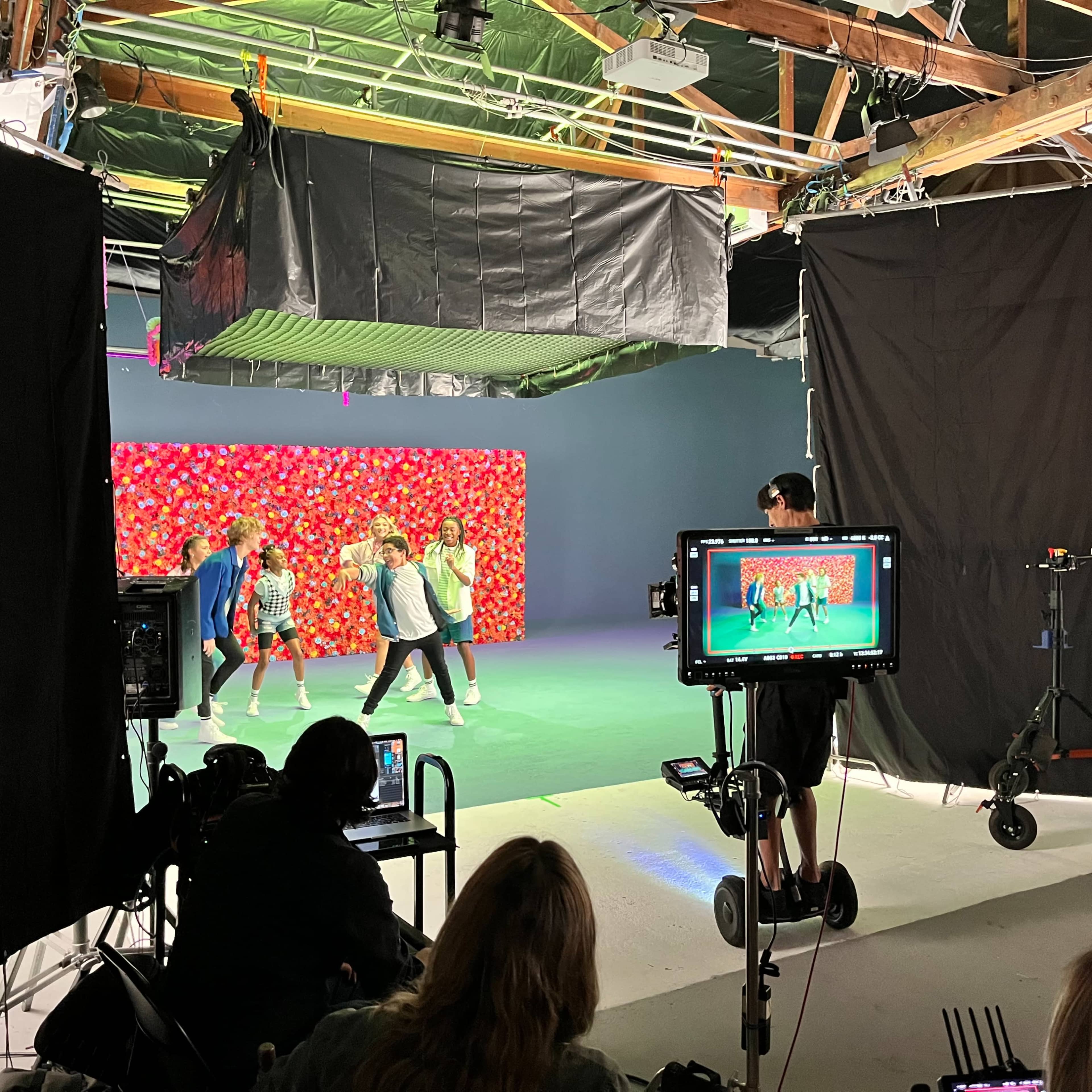 A filming studio with a colorful backdrop, where a group of dancers is performing while a crew member operates a camera on a tripod.