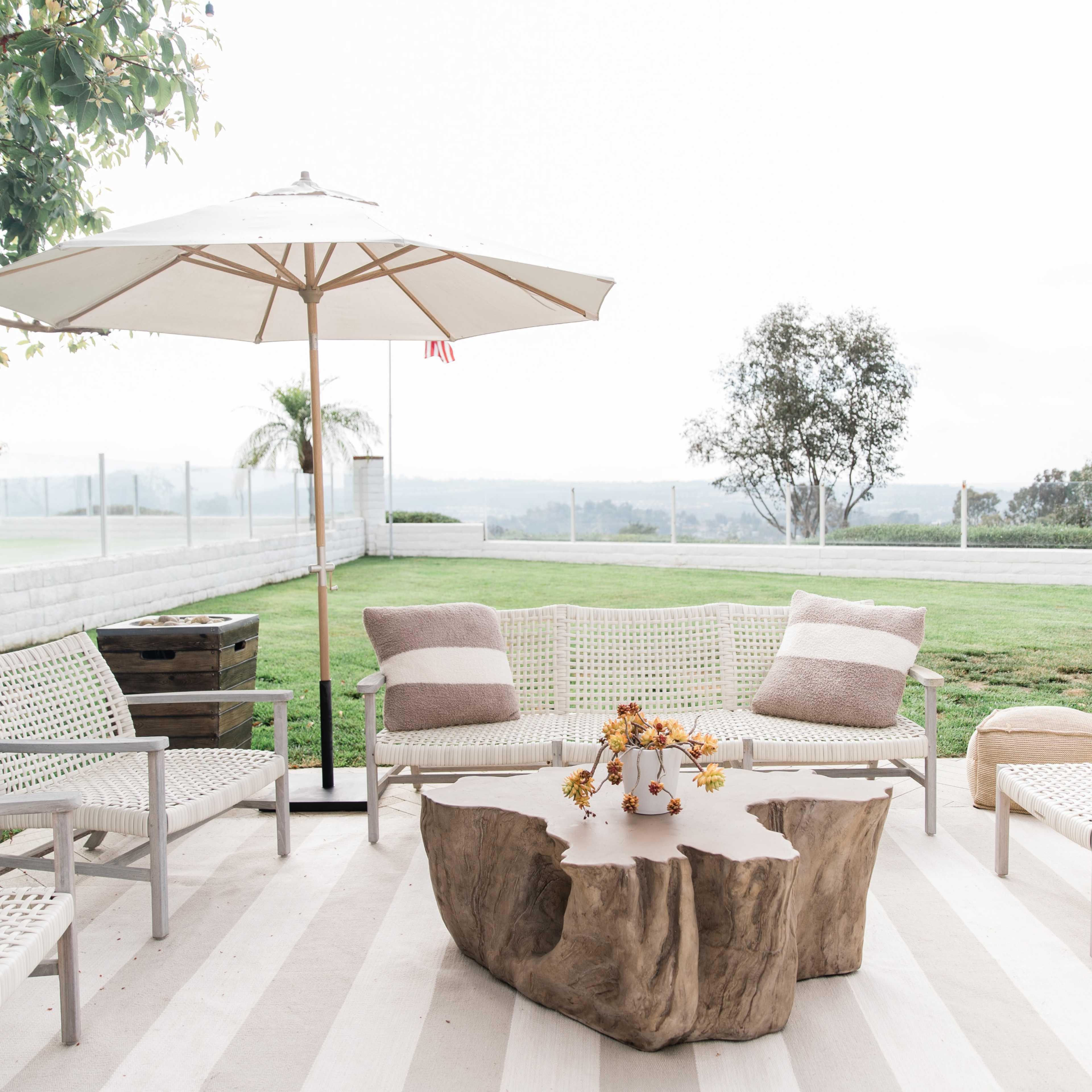 A patio area features a wooden coffee table surrounded by several chairs and a large umbrella, with a grassy landscape visible in the background.