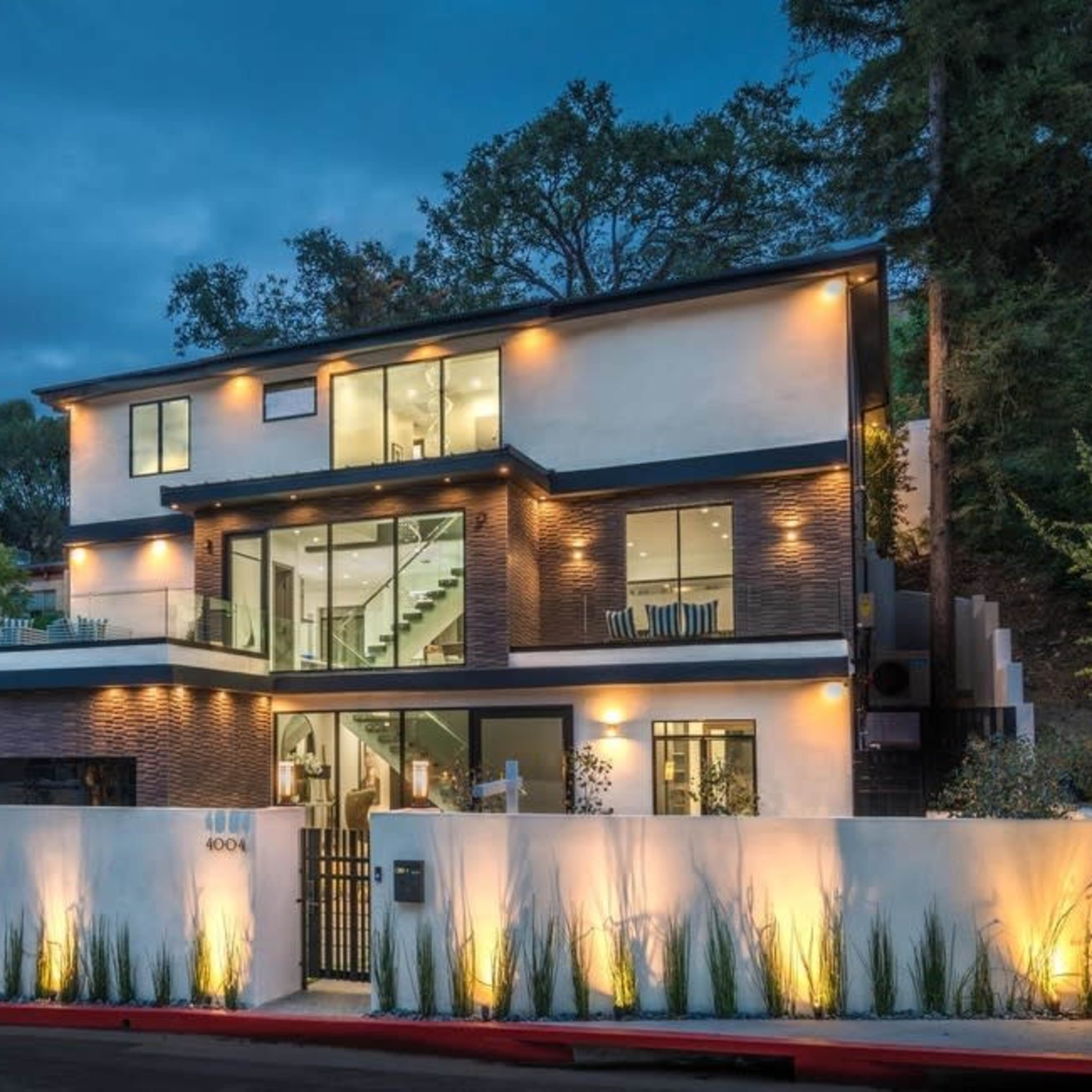 A modern, three-story house with large windows and a sleek design stands illuminated at dusk, surrounded by greenery and featuring a gated frontyard.