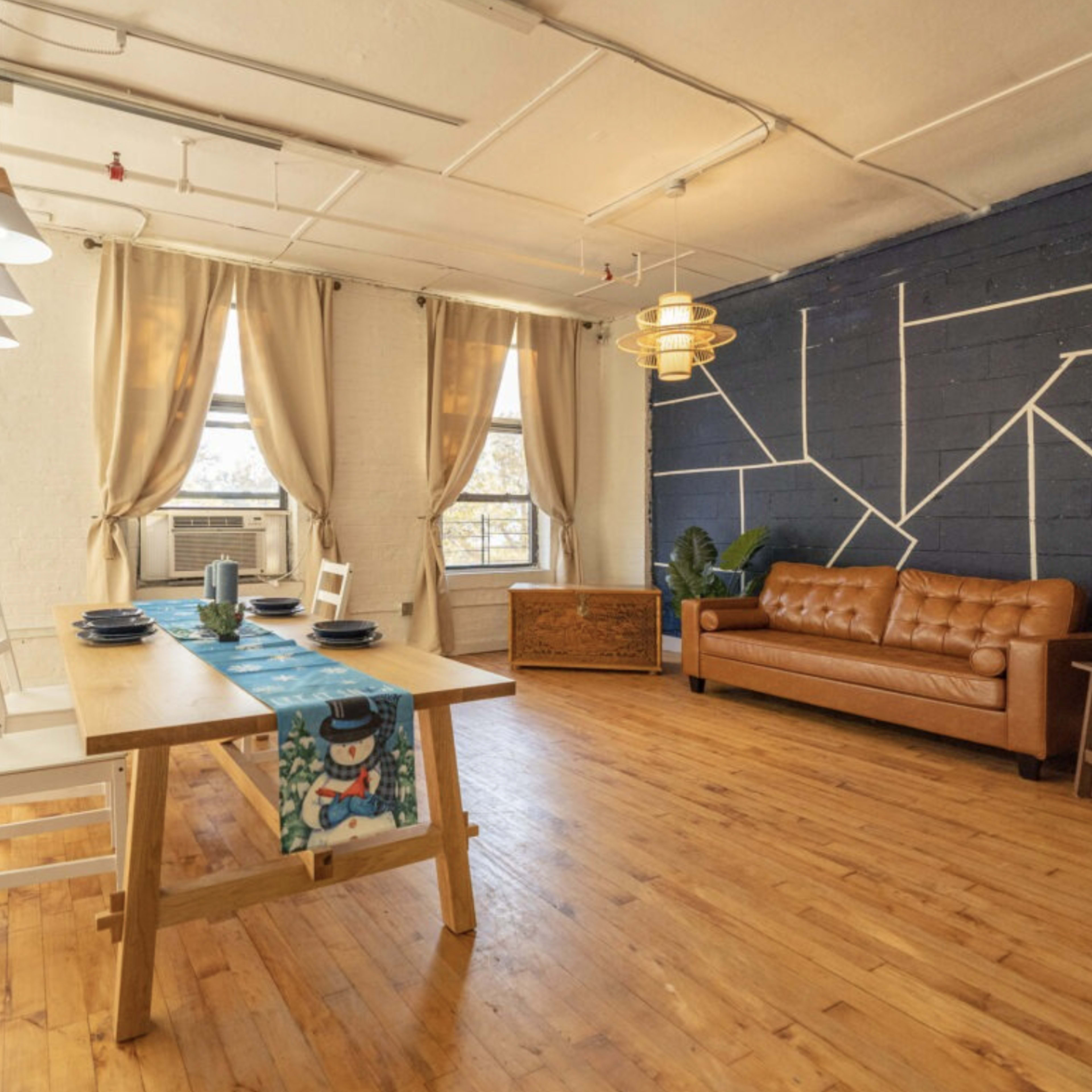A spacious room featuring a wooden dining table with chairs, a brown leather sofa, and large windows, accented by a blue wall with geometric white lines.
