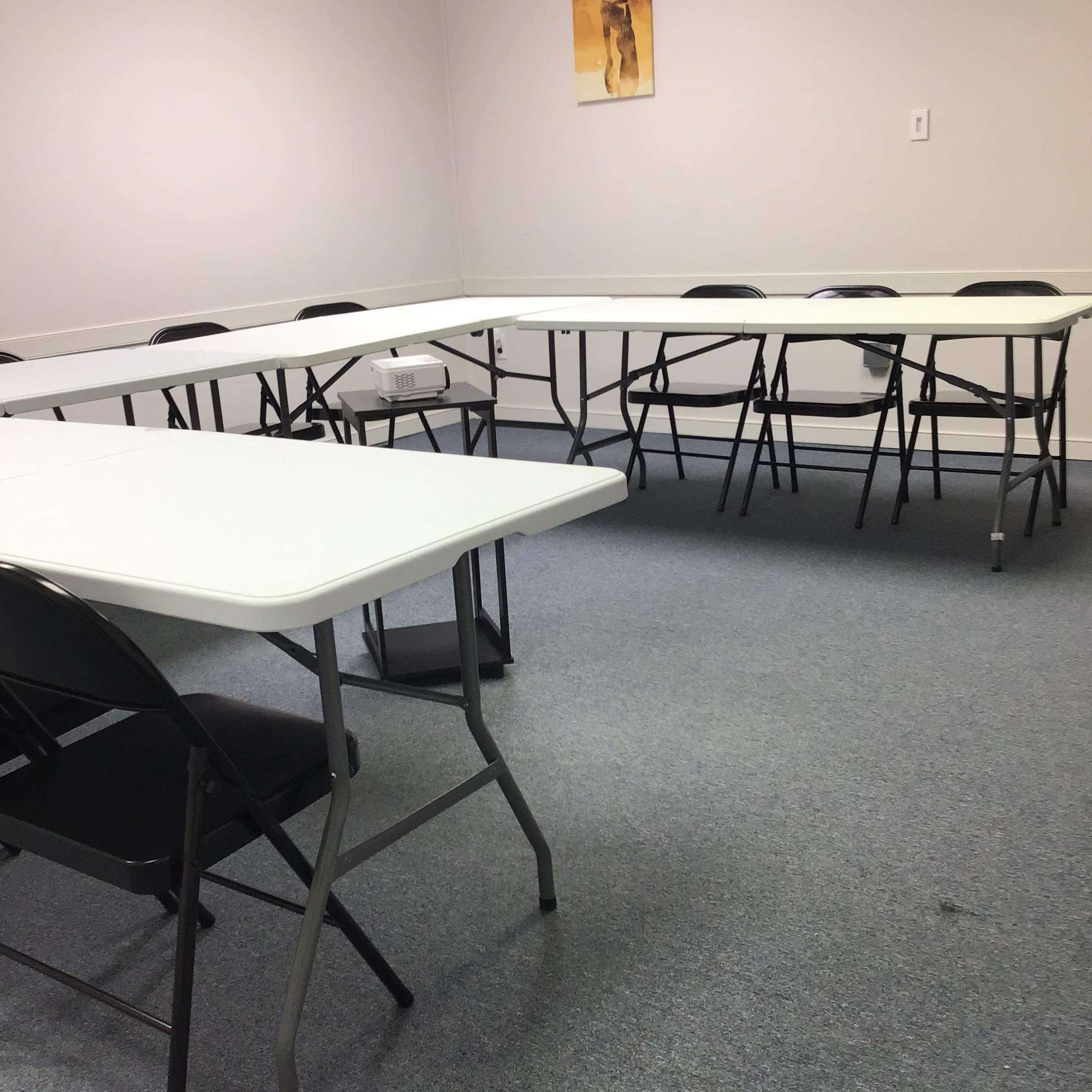 A rectangular room is set with several folding tables arranged in a U-shape, surrounded by black chairs on a carpeted floor.